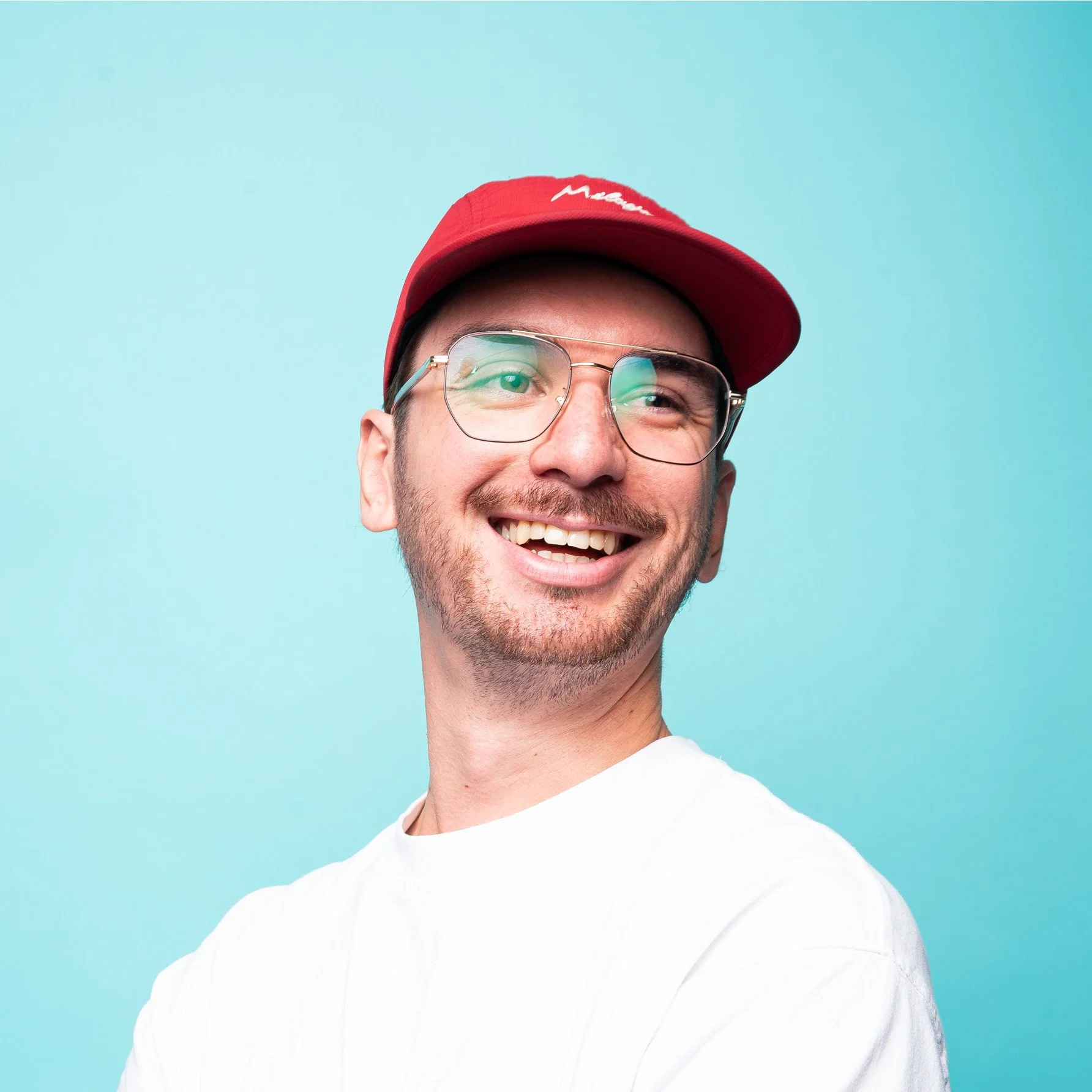 A smiling man wearing glasses, a red cap, and a white shirt against a light blue background.