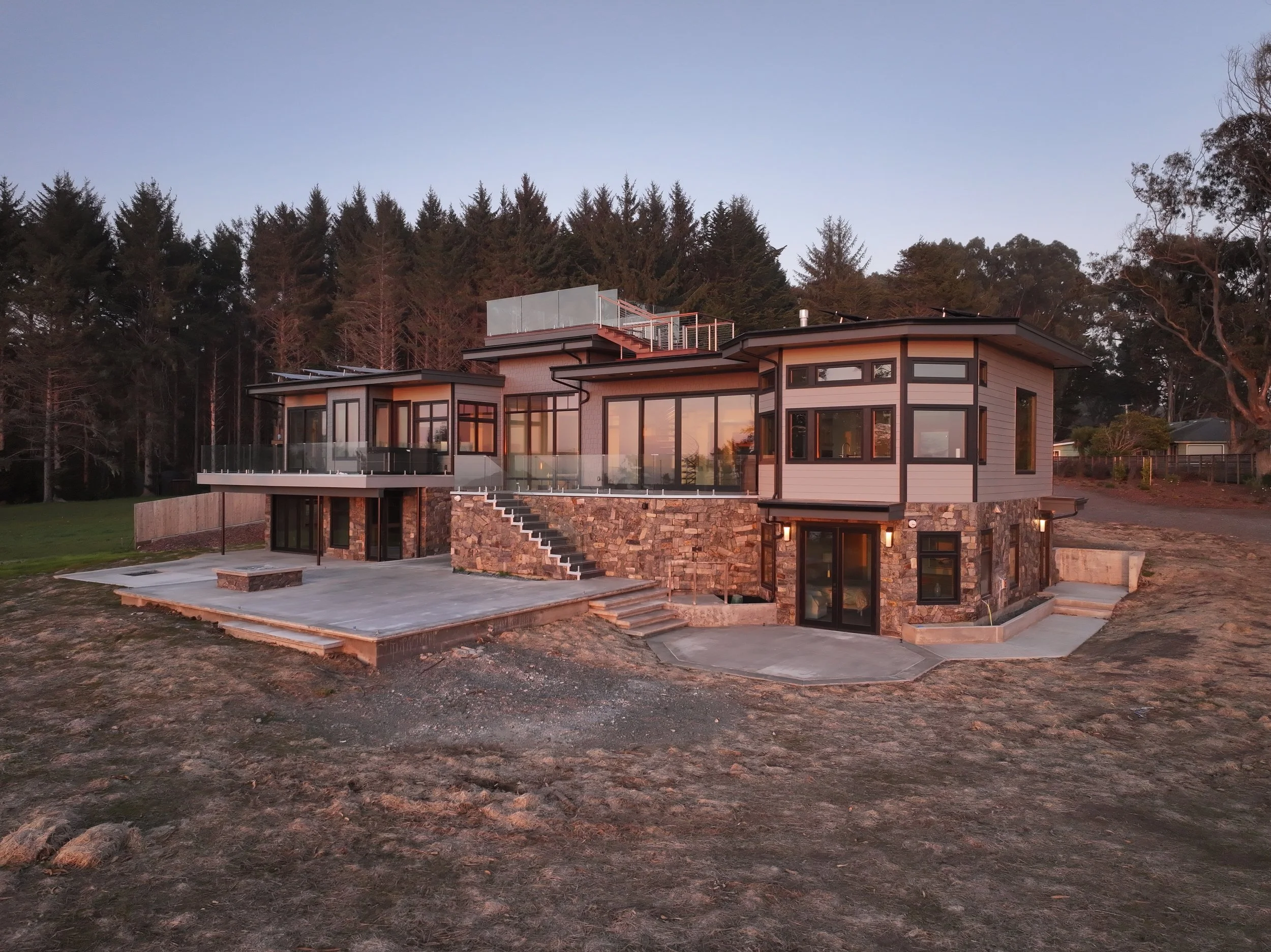 Modern multi-story house with large windows, stone and siding exterior, situated in a semi-rural area with trees in the background at sunset.
