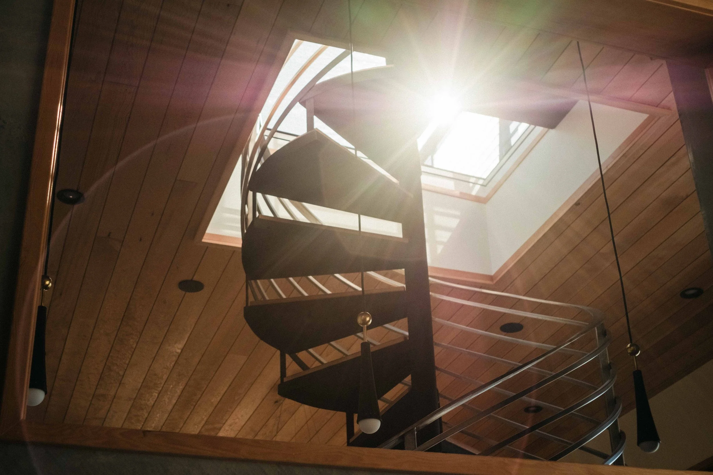 Interior view of a wooden staircase with spiral design, sunlight coming through a skylight, and modern black pendant lights hanging from the ceiling.