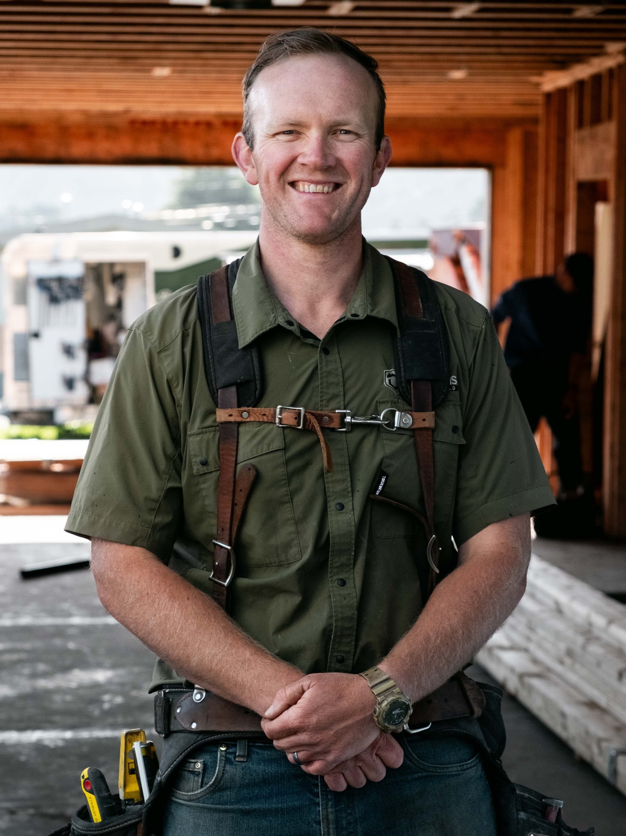 A smiling man wearing a green shirt and a watch, standing indoors with a wooden ceiling and some people in the background.