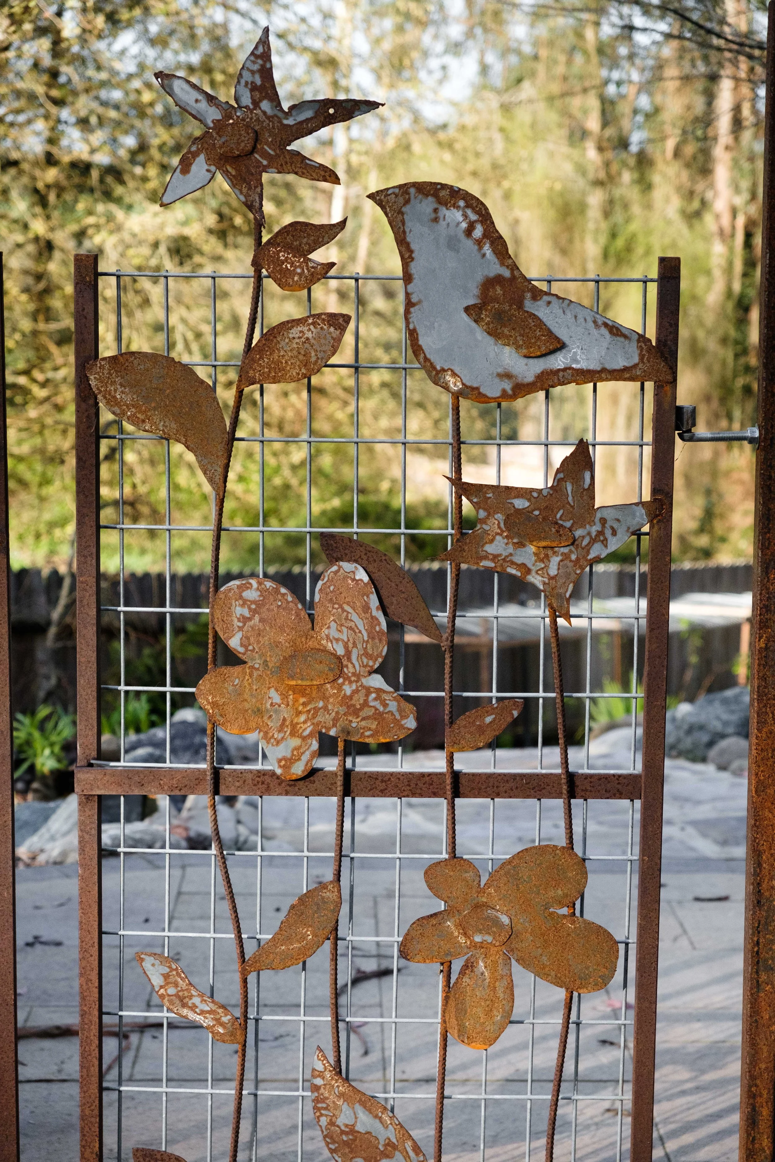 Rusty metal garden art with flowers, leaves, and birds on a wire fence, outdoors with trees and a patio in the background.
