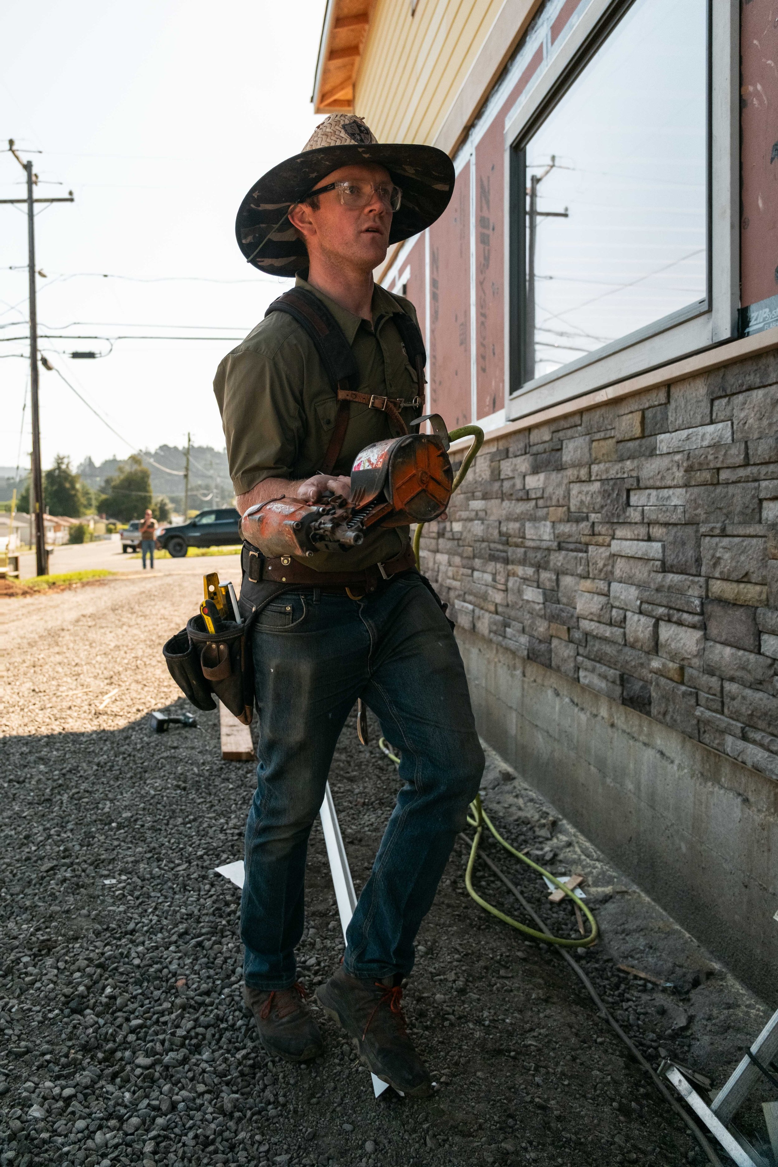 A construction worker wearing a large hat, safety glasses, and a tool belt, walking near a building with stone siding while holding a power tool.