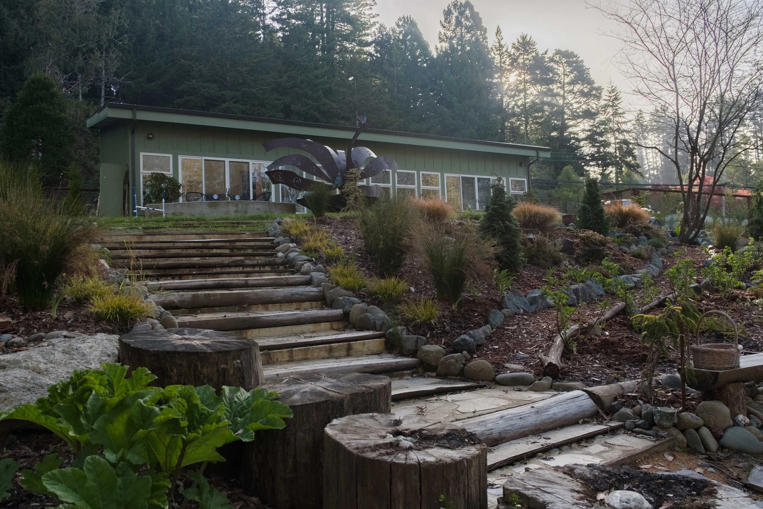 A landscaped garden with wooden stairs leading up to a green house with large windows, surrounded by trees and plants, with a metal butterfly sculpture near the house.