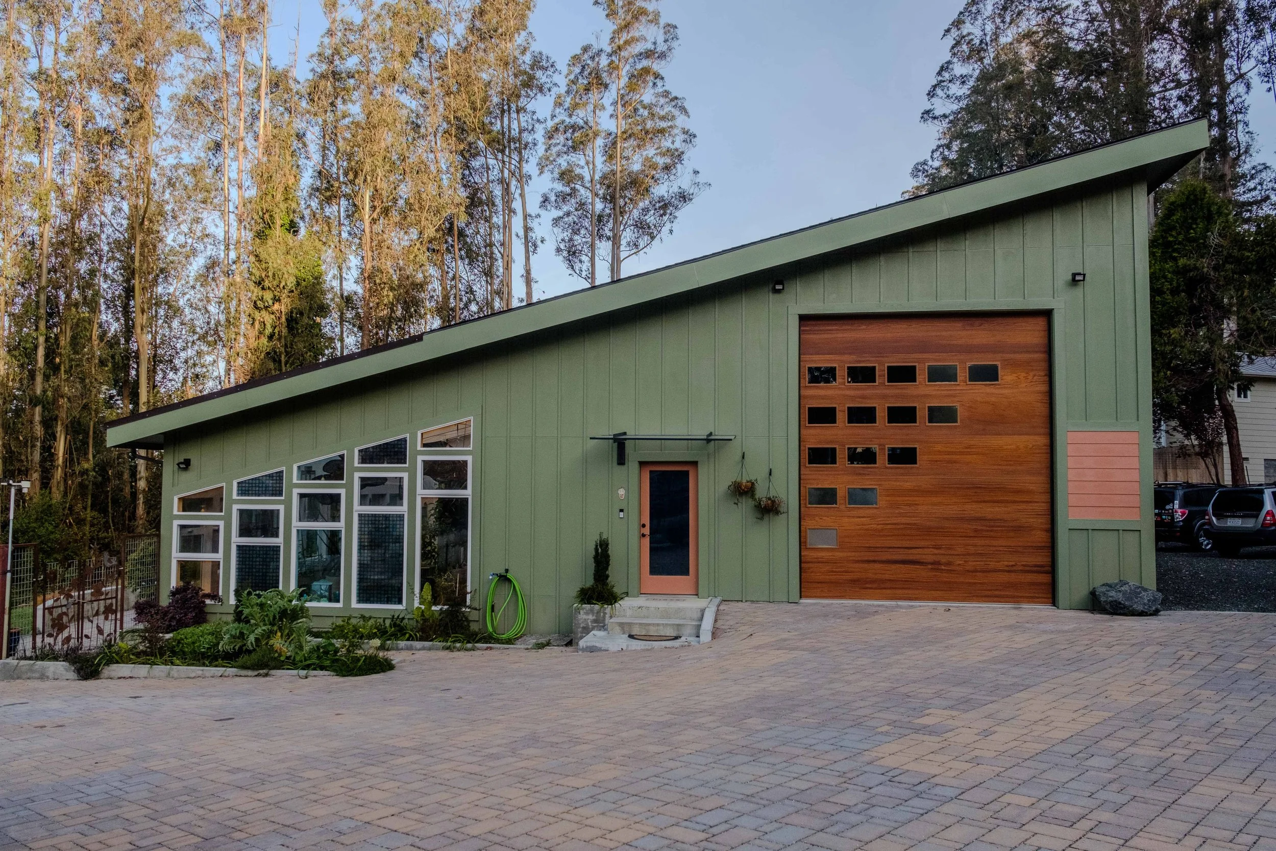 Modern house with a green exterior, large windows on the left side, and a wooden garage door on the right side, set against a backdrop of tall trees.