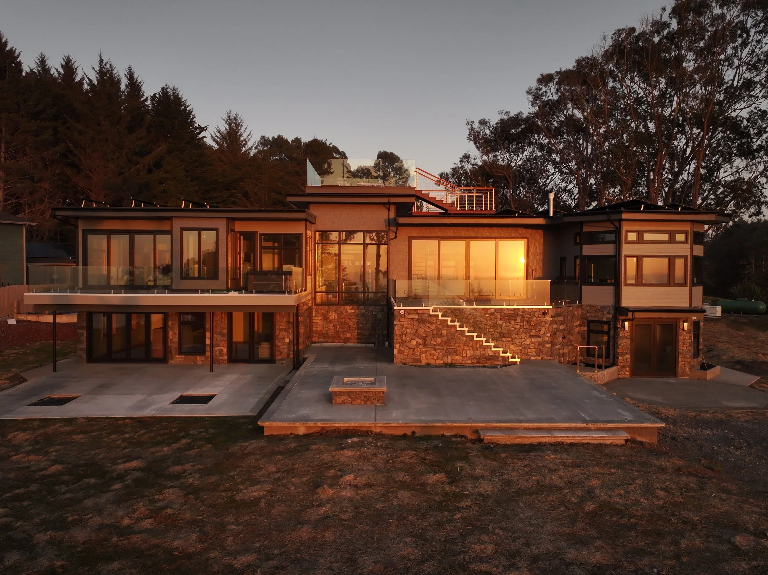 Modern house with large glass windows and stone accents, set against a backdrop of trees at sunset.