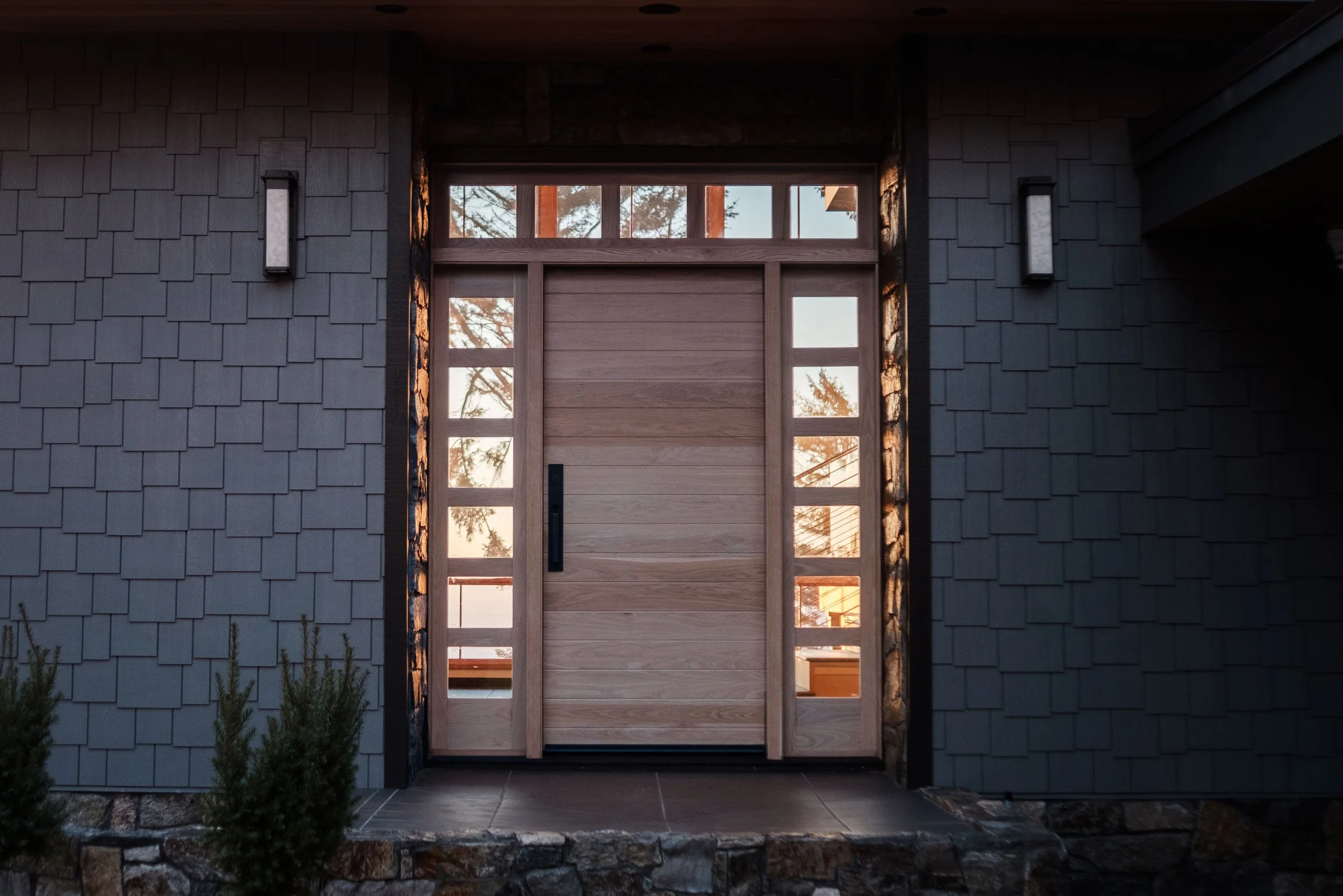 Front entrance of a modern house with a wooden door and windows, dark exterior walls, and outdoor lighting