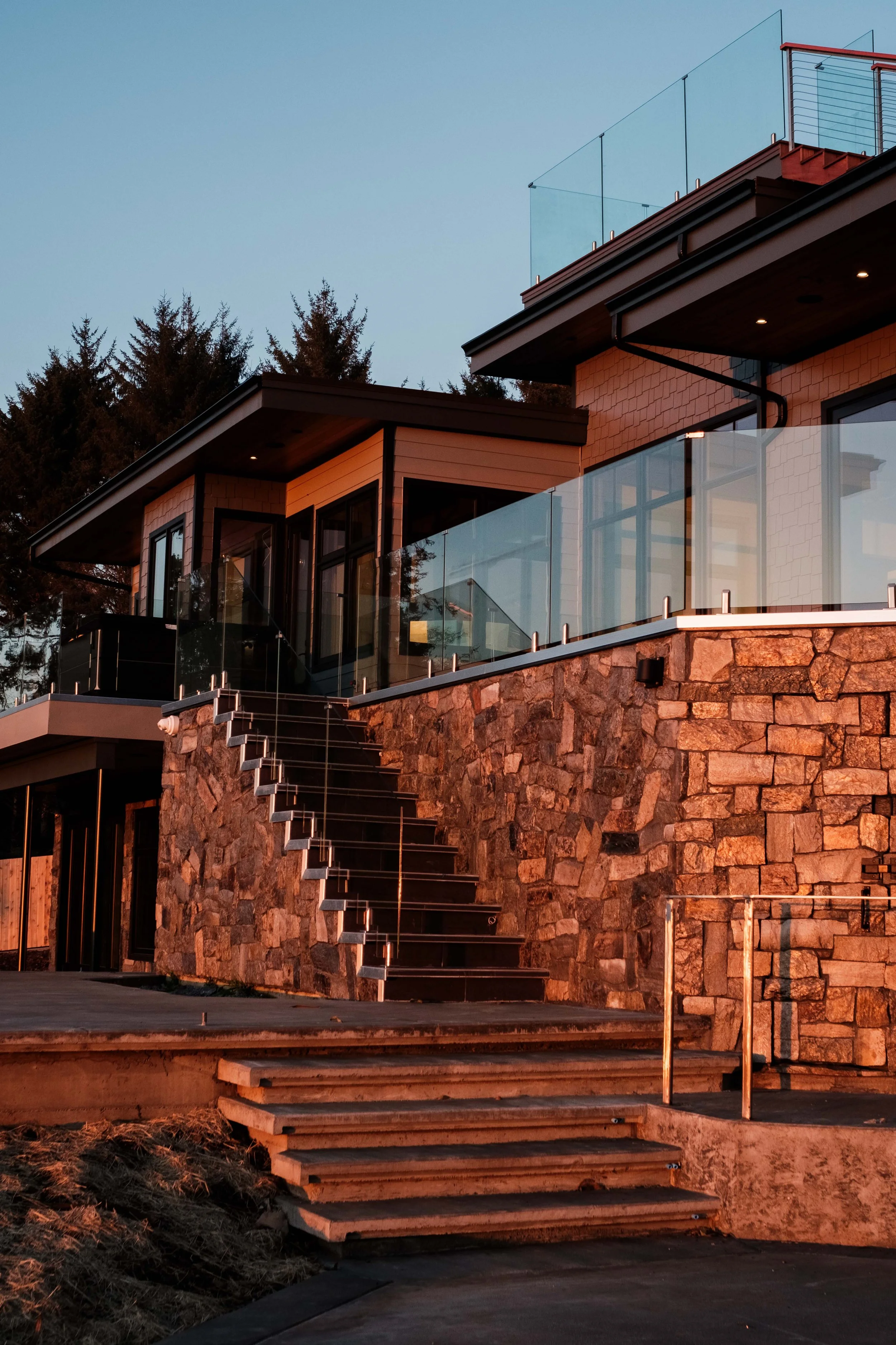 Modern multi-level house with stone steps, glass railings, large windows, and a balcony, during sunset.