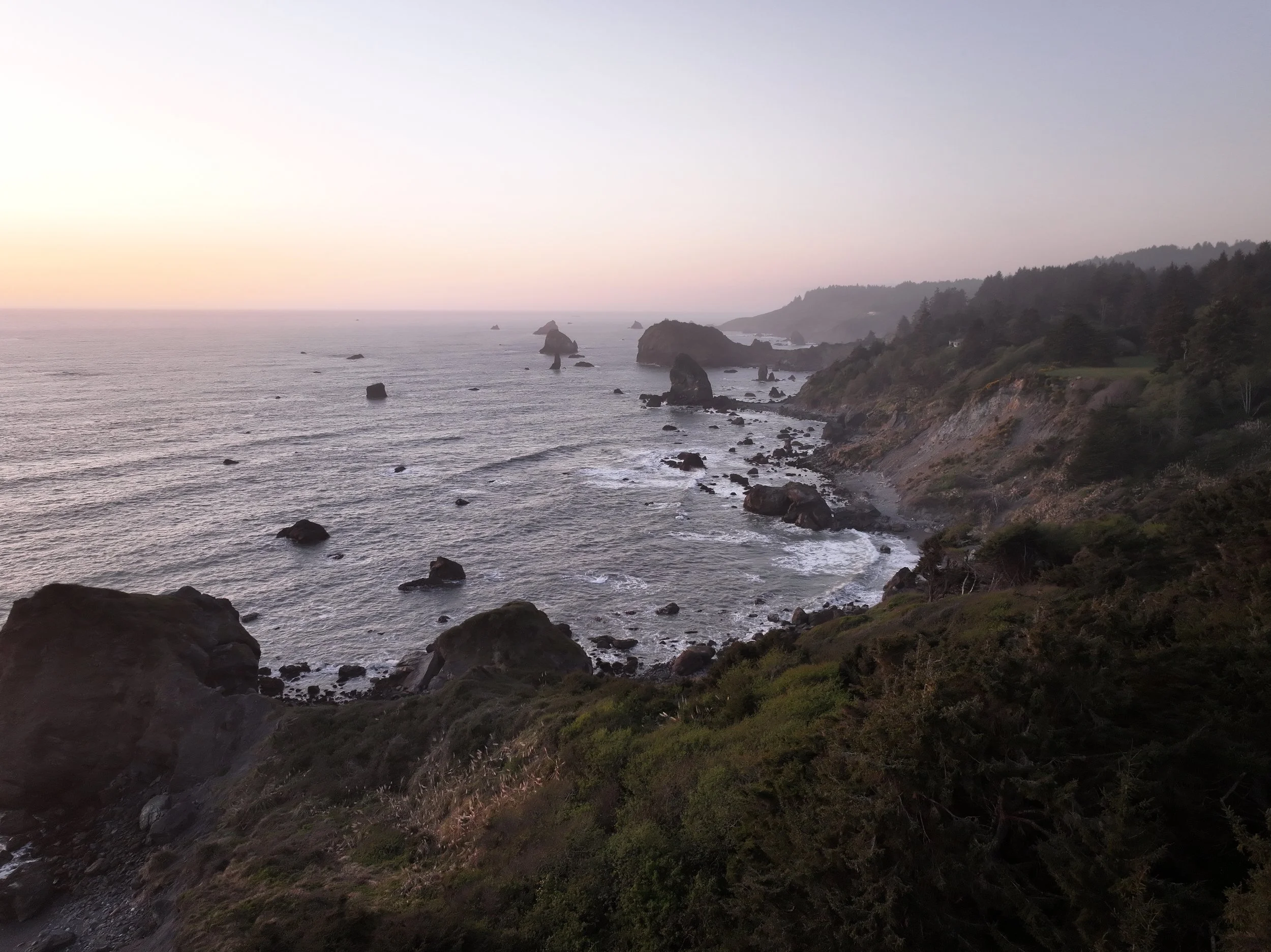 A rugged coastline at sunset with large rocks in the ocean, steep cliffs with greenery, and a distant view of the sea and sky.