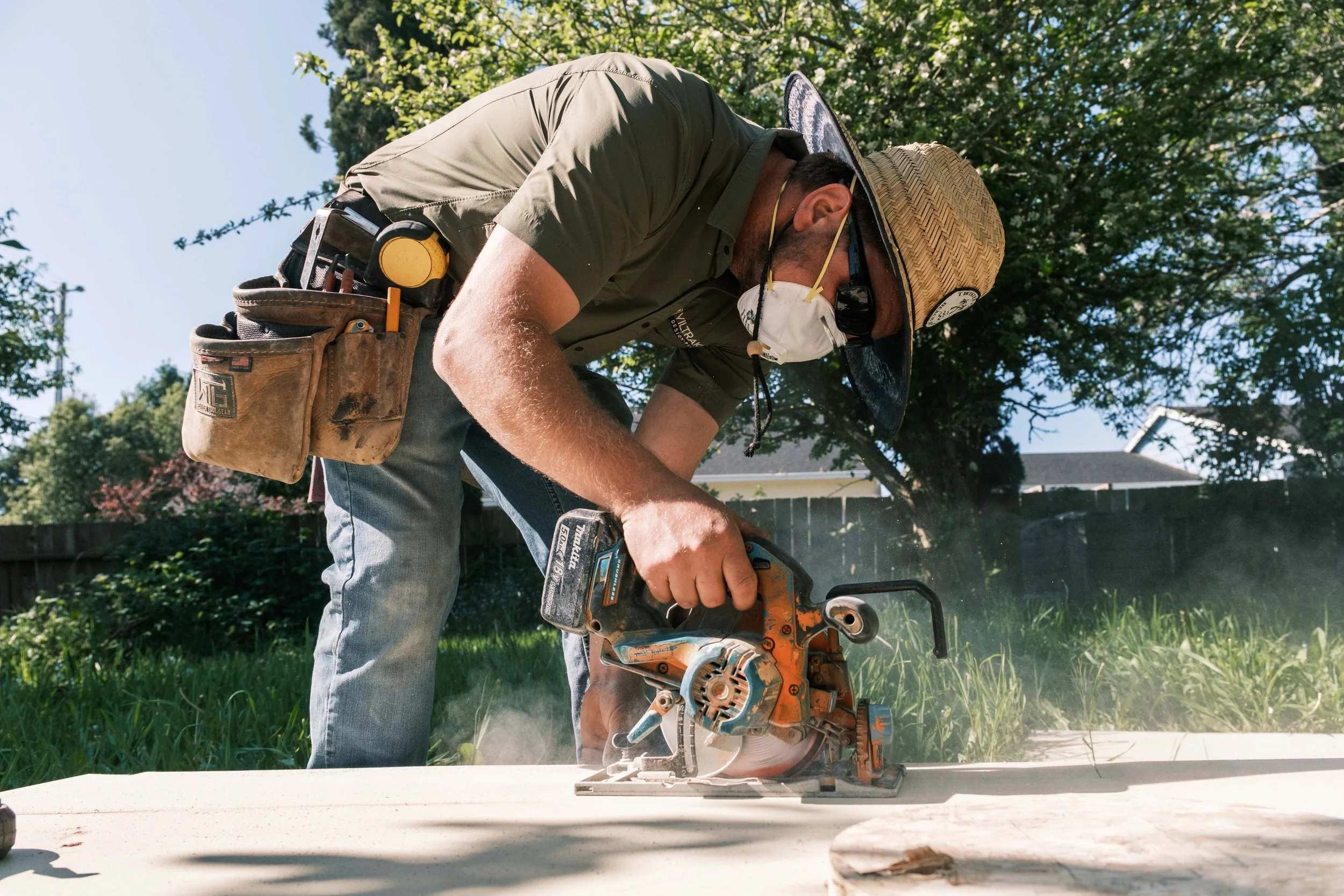 Man wearing a straw hat, sunglasses, and a face mask using a circular saw to cut wood outdoors on a sunny day.