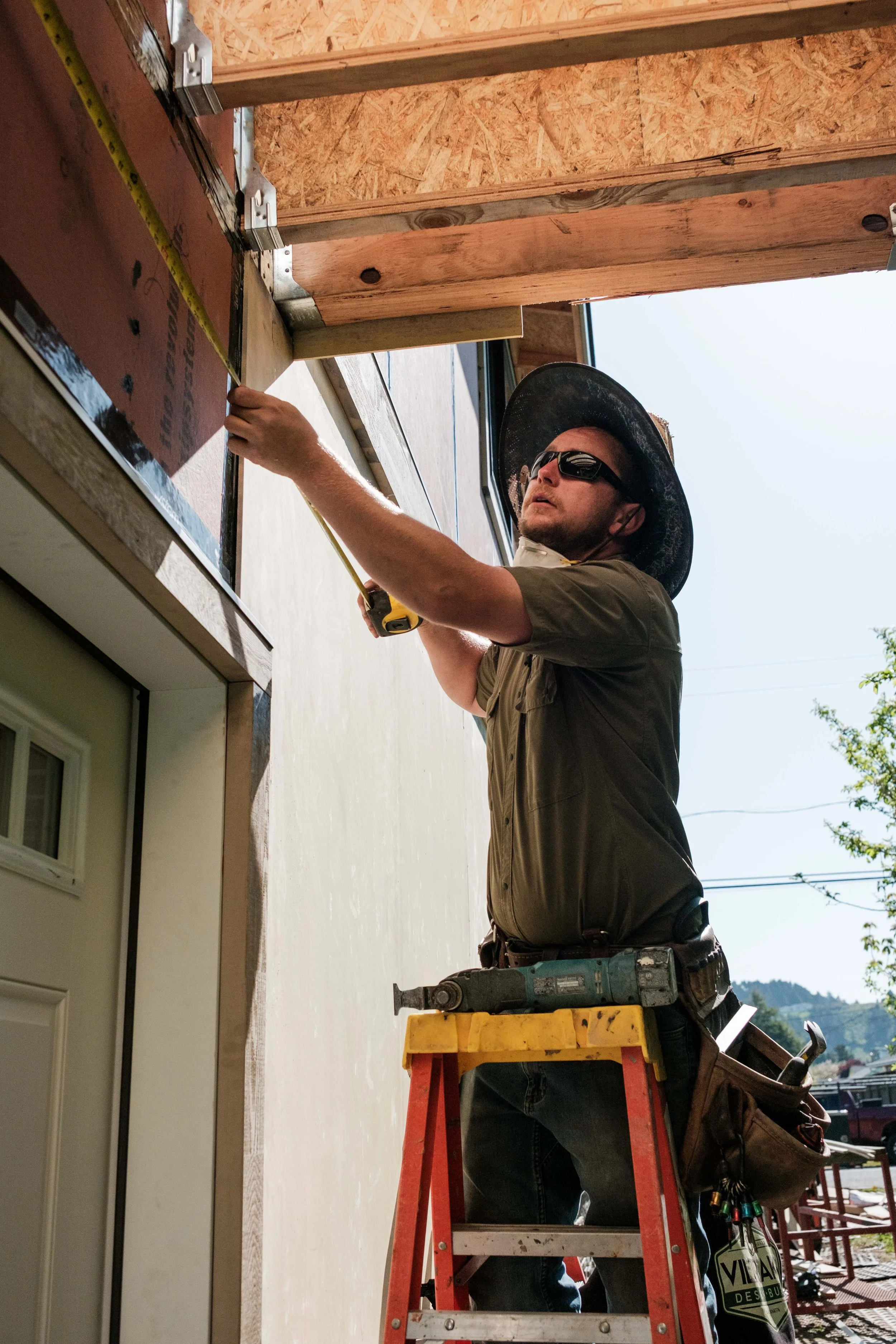A construction worker measuring the side of a building while standing on a ladder. He is wearing sunglasses, a wide-brimmed hat, a tool belt, and a dark green shirt. A power sander rests on the ladder beside him.