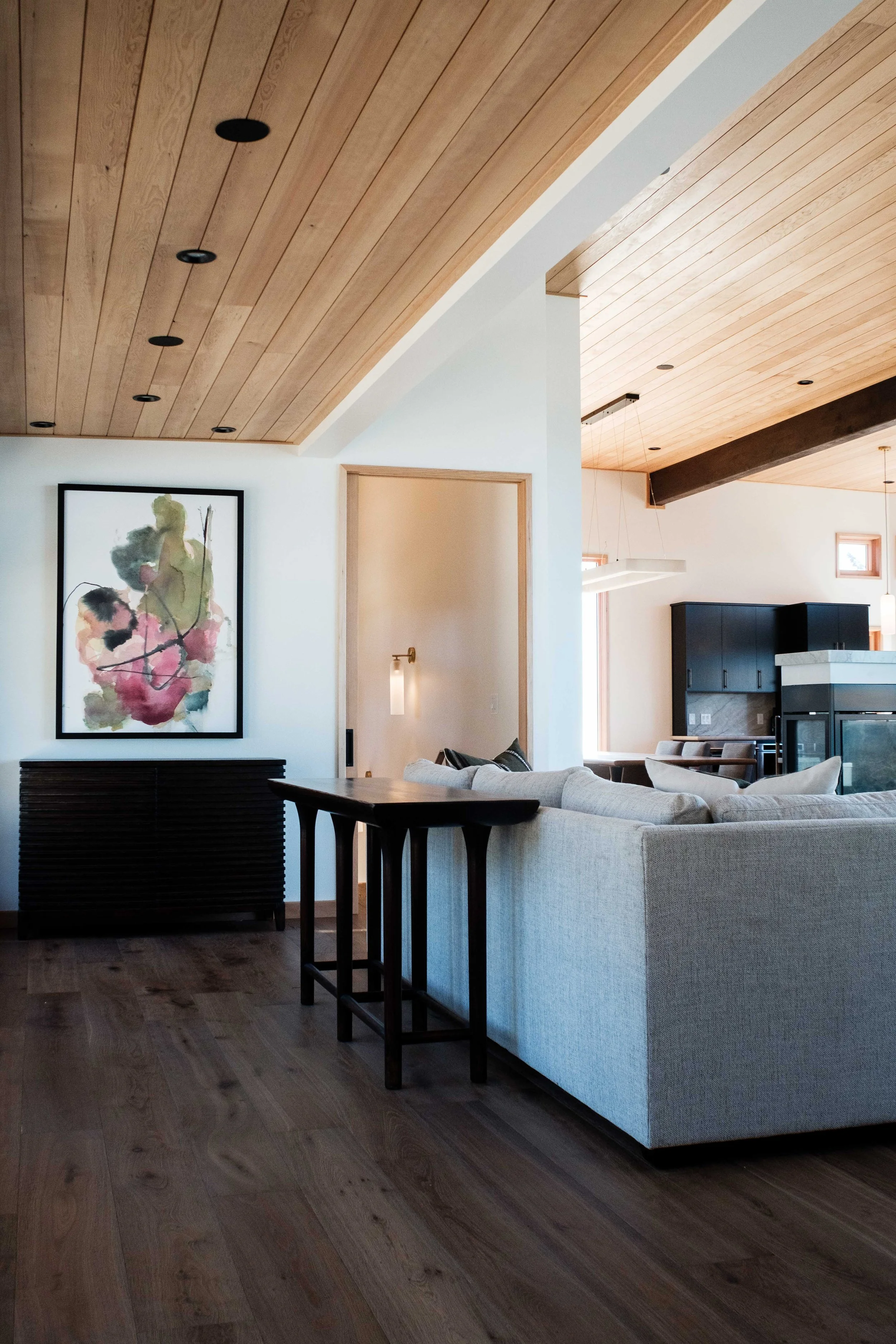 Modern living room with a beige sofa, wooden ceiling, wooden flooring, abstract art on the wall, and part of the kitchen in the background.