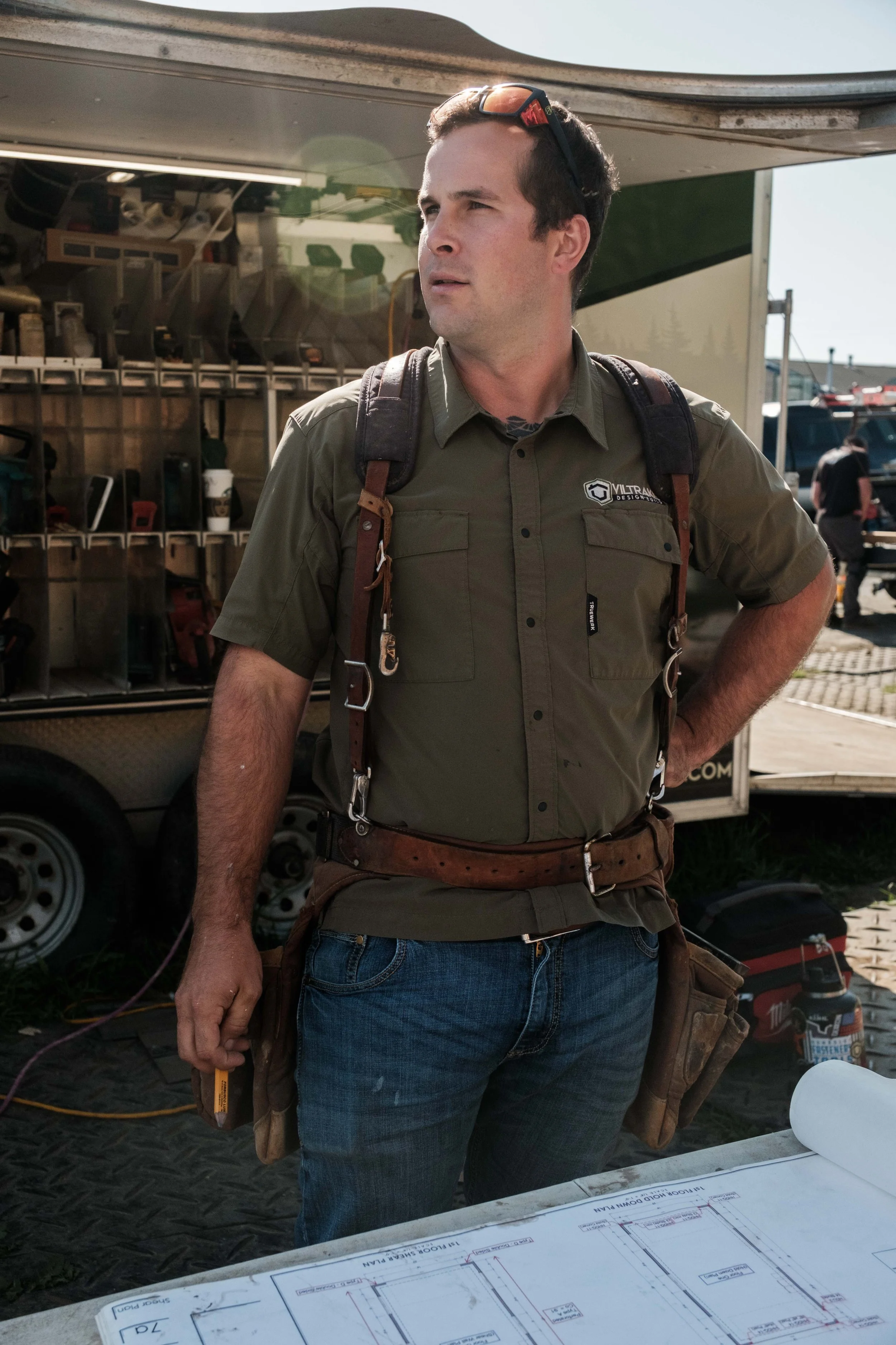 A man in a green work shirt with a logo, standing outdoors in front of trailers and equipment, with a large blueprint on a table in front of him.