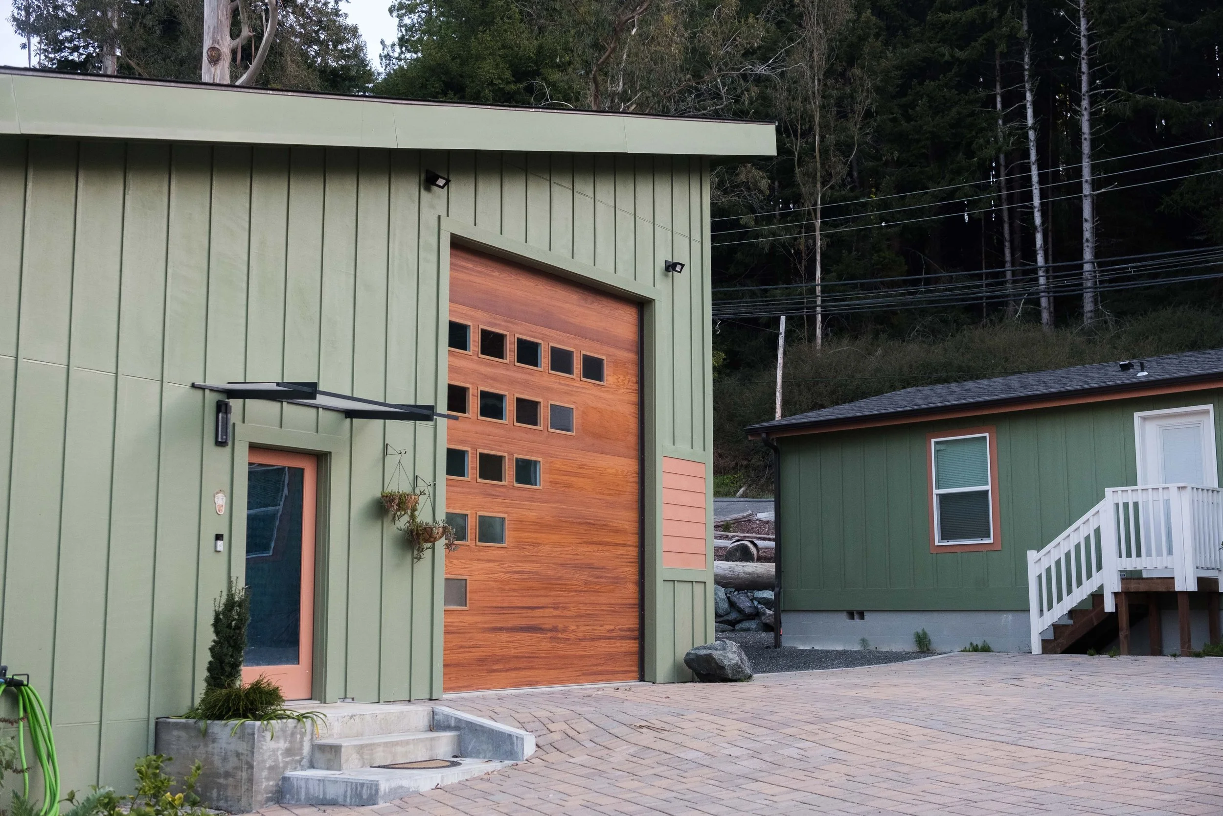 Photo of a green modern house with a large wooden garage door, adjacent to a smaller green building with a white staircase, surrounded by trees and a paved driveway.