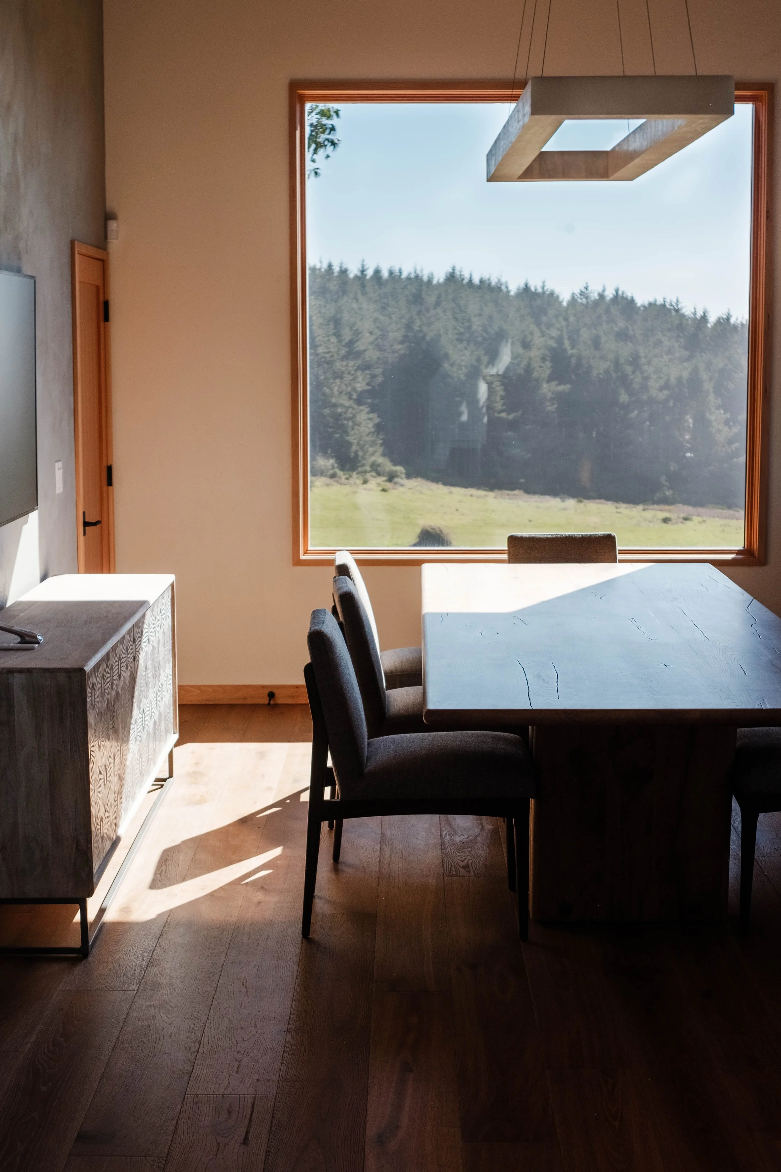 A dining room with a large window showing a view of trees and greenery outside. There is a wooden table, six dark-colored chairs, a television on a sideboard, and a modern rectangular light fixture hanging from the ceiling.