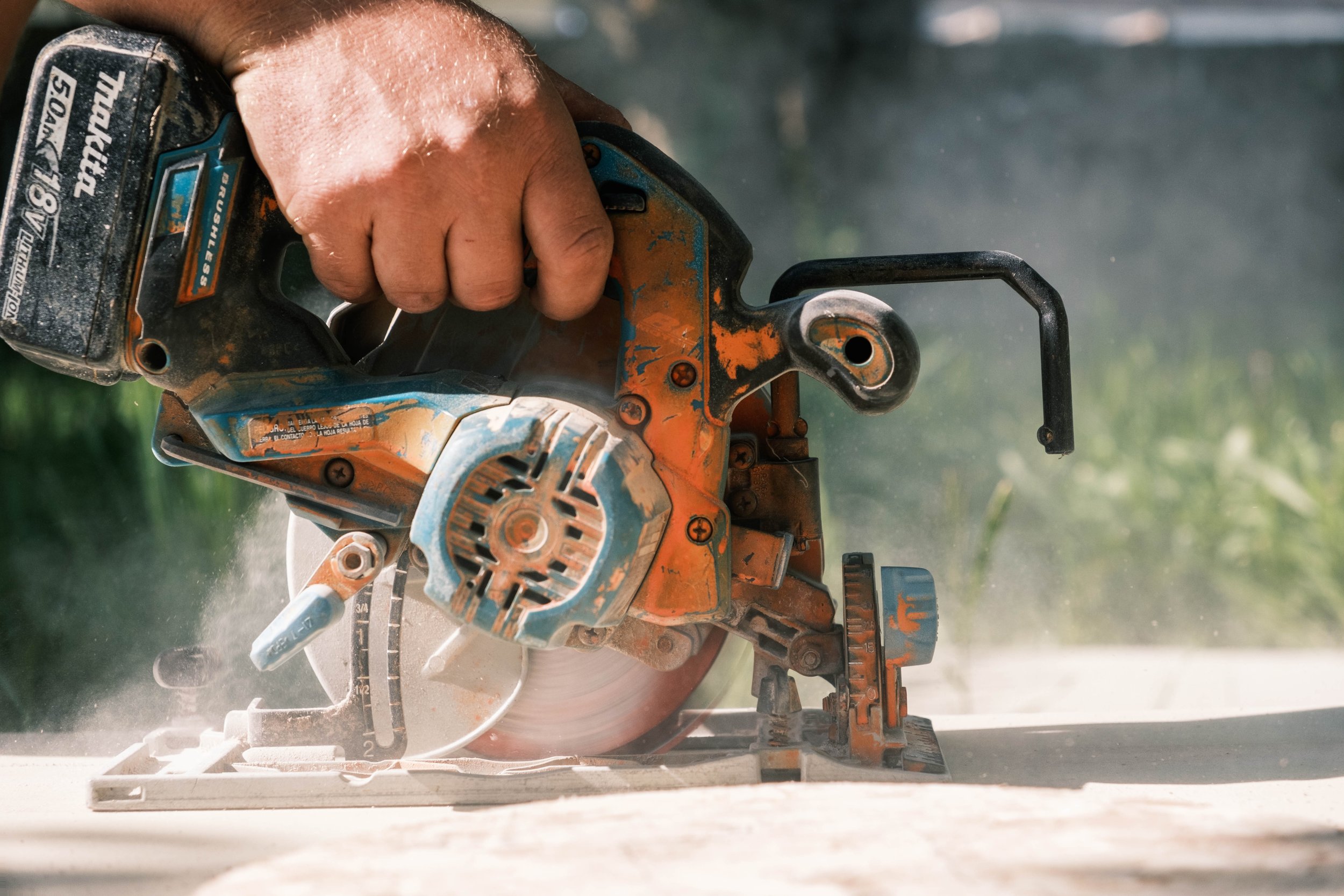 A person's hand operating an orange and blue circular saw cutting through wood outdoors, with dust and wood particles flying.