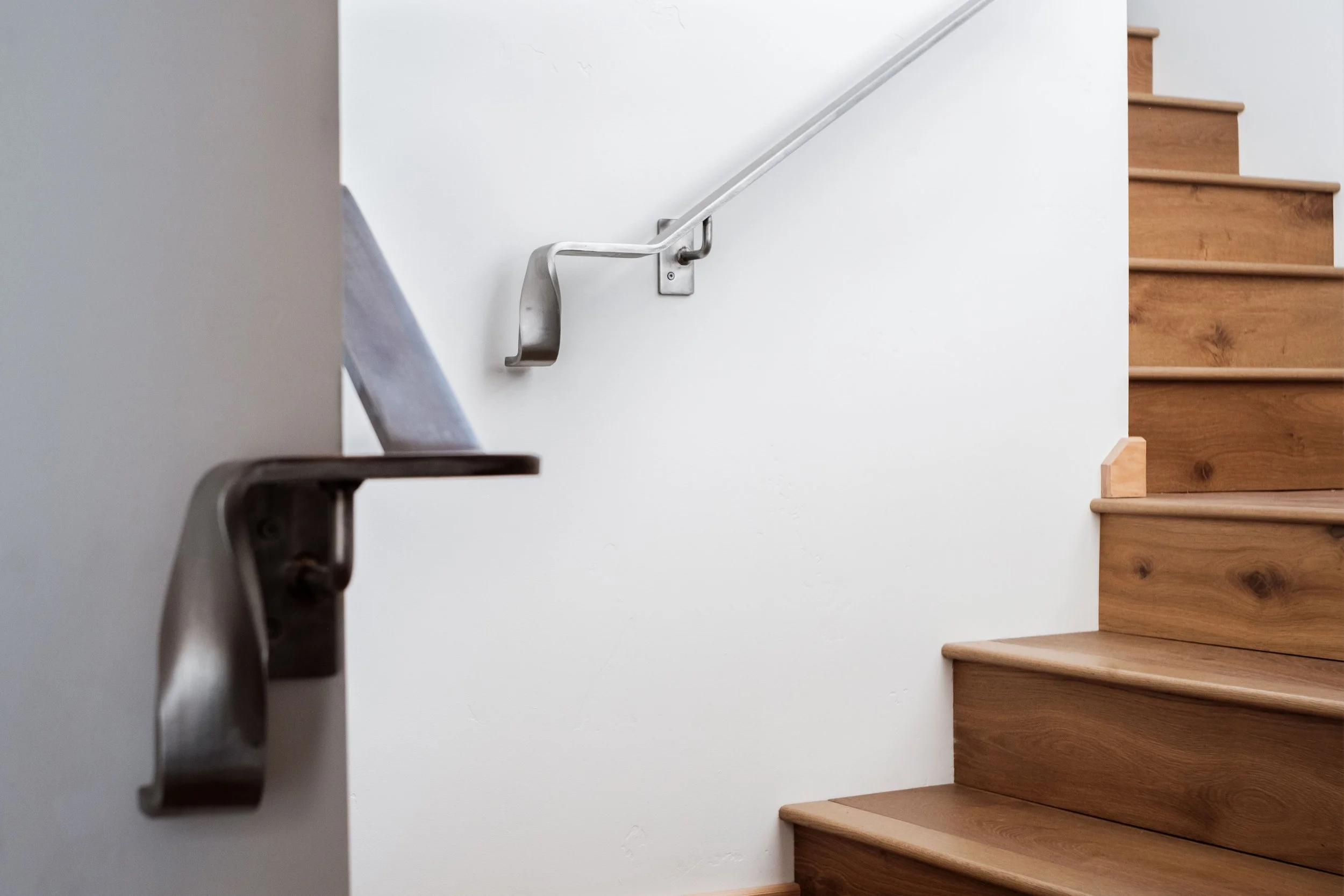 Wooden stairs with a metal handrail on a white wall, viewed from the bottom of the staircase.