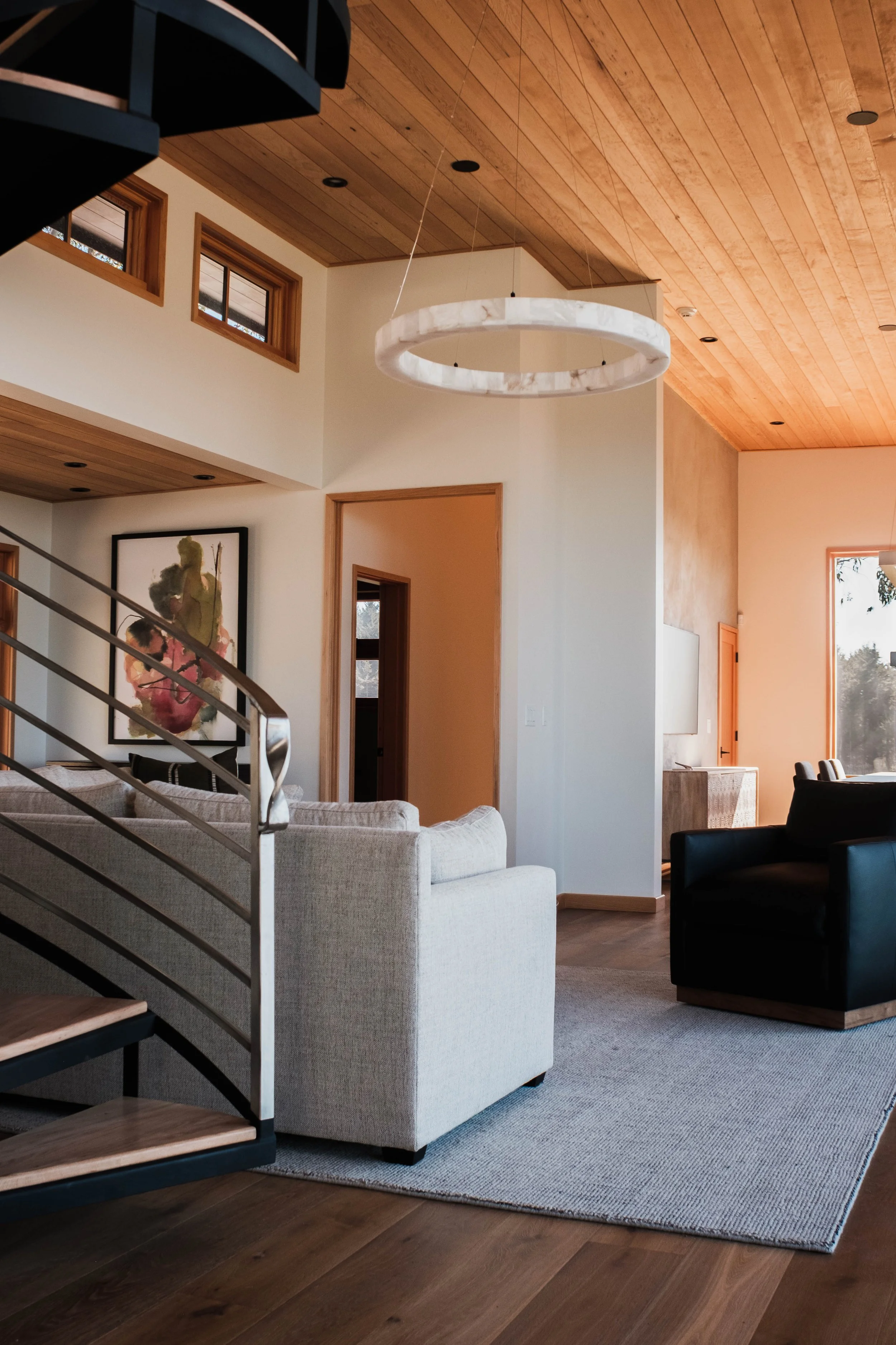 Living room with wooden ceiling, Modern circular chandelier, beige sofa, black armchair, artwork on wall, windows letting in natural light, wooden flooring, area rug, staircase railing.