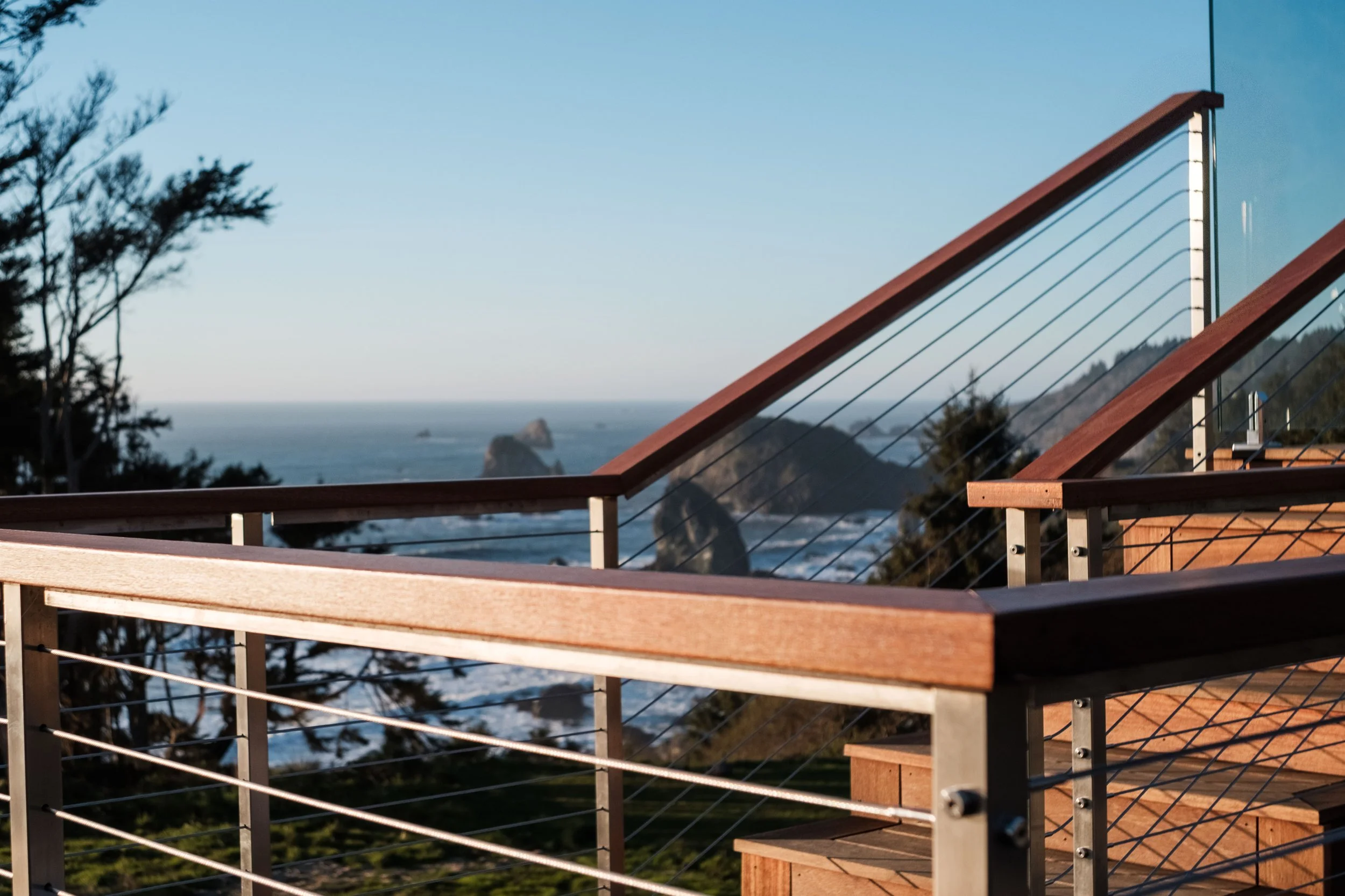 View of a wooden deck railing overlooking a coastal landscape with ocean, large rocks, and hills in the distance, on a clear day.