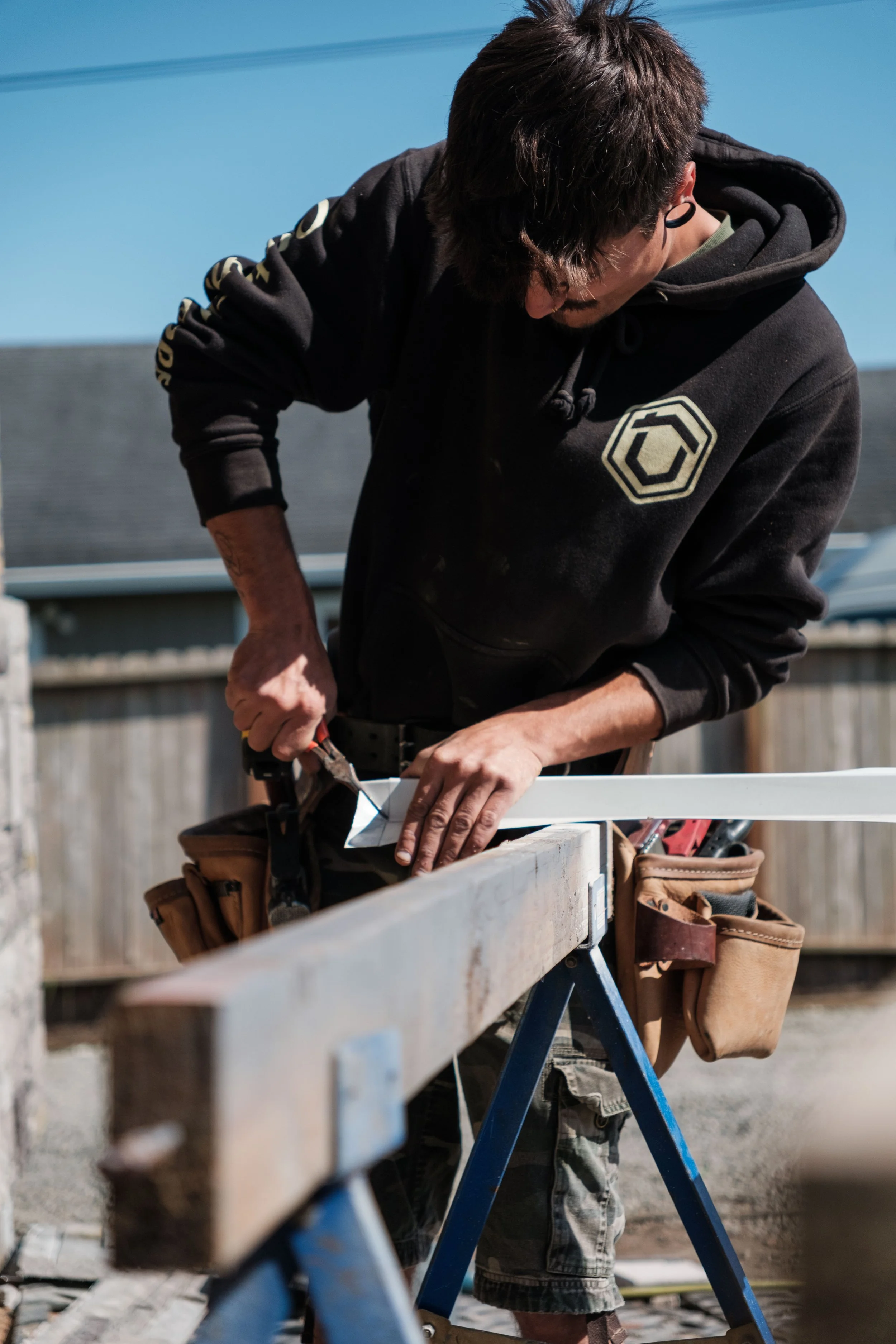 A man working on woodworking outdoors, measuring a piece of wood with a speed square on a sawhorse.