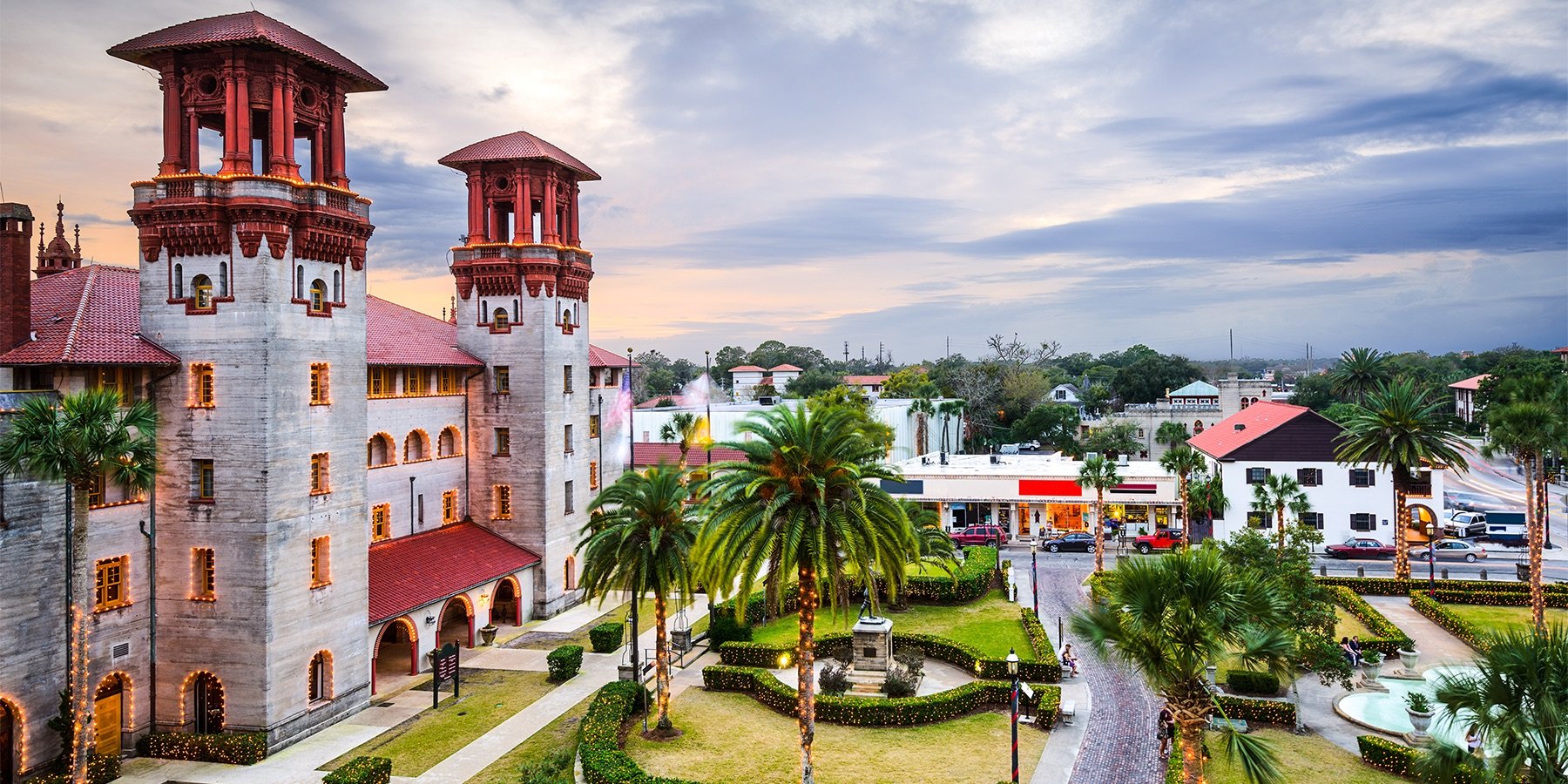 A scenic view of a historic castle-like building with white walls, red-tiled roofs, and tall towers, surrounded by palm trees and a well-maintained park at sunset.