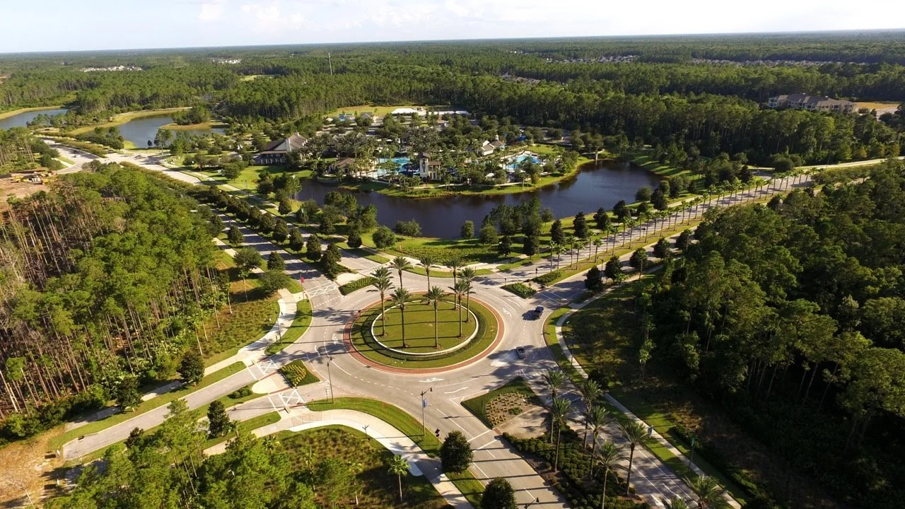 An aerial view of a landscaped park with a roundabout featuring palm trees, a small body of water, walking paths, and green trees surrounding the area.