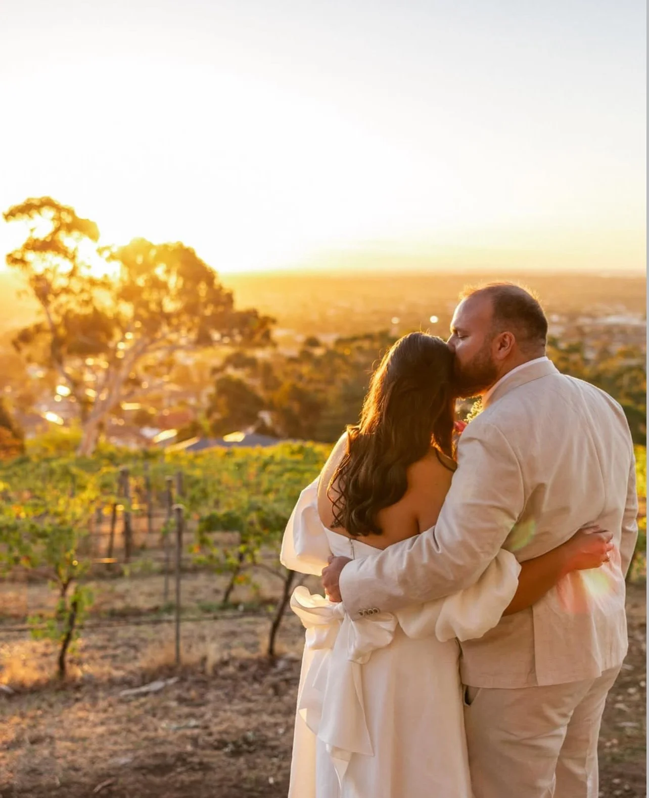 woodfolk wedding couple with live music Adelaide. A couple sharing a kiss during sunset in a vineyard, dressed in white wedding attire.