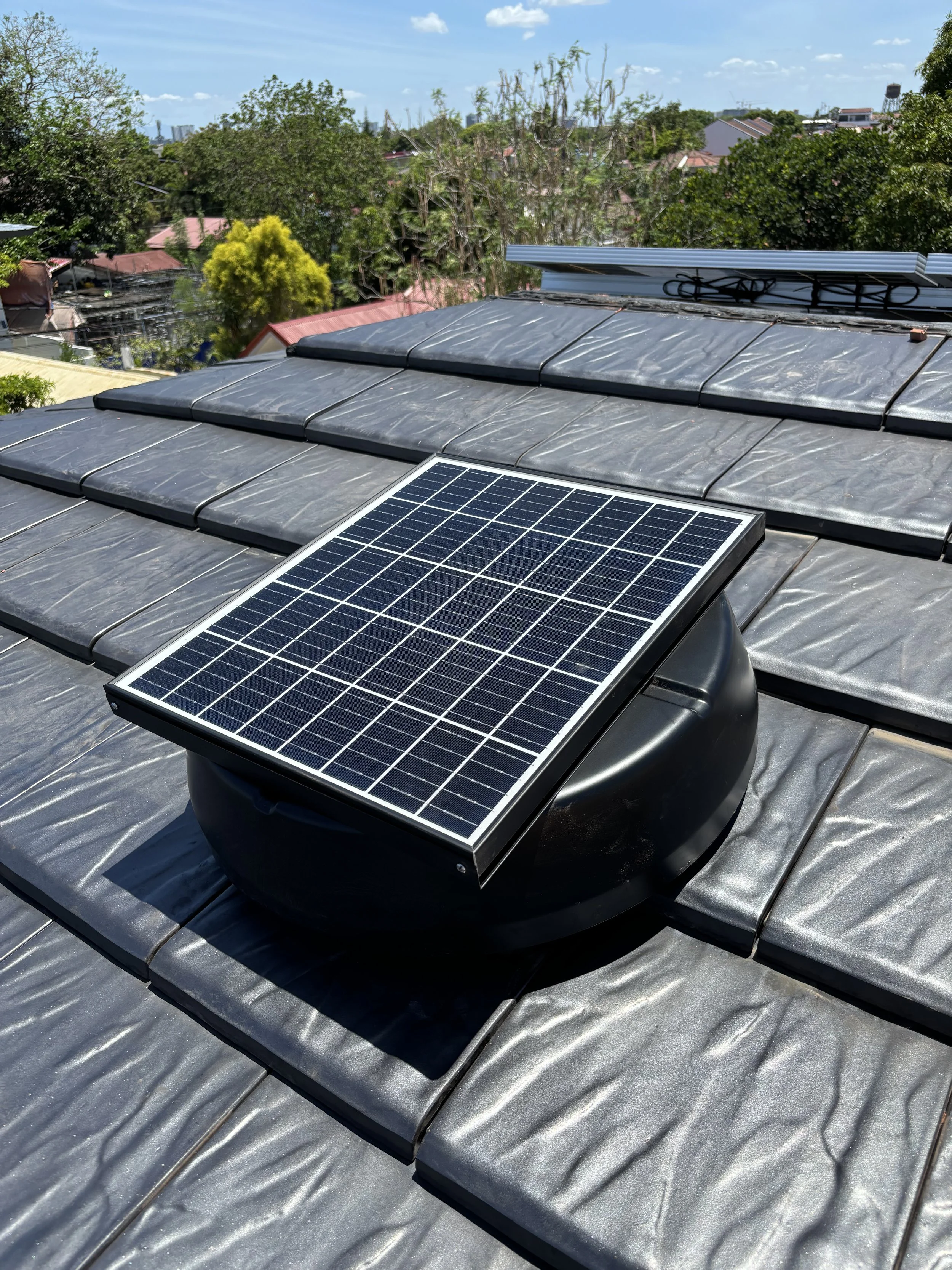 A solar-powered exhaust fan installed on a metal roof, with rooftops and trees in the background under a partly cloudy sky.