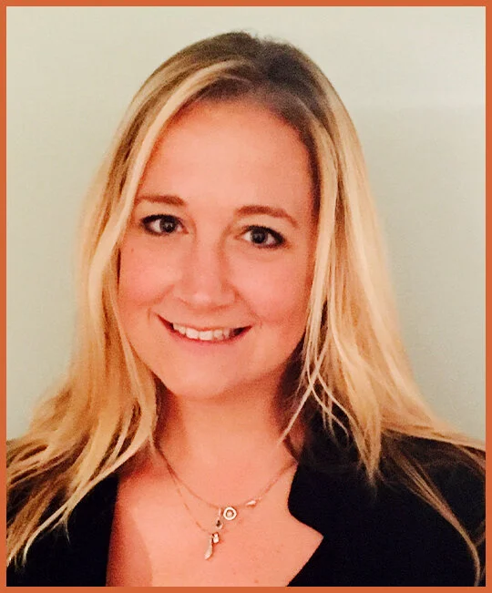 A woman with long blonde hair smiling at the camera, wearing a black top and a gold necklace with pendants, against a plain light-colored background.