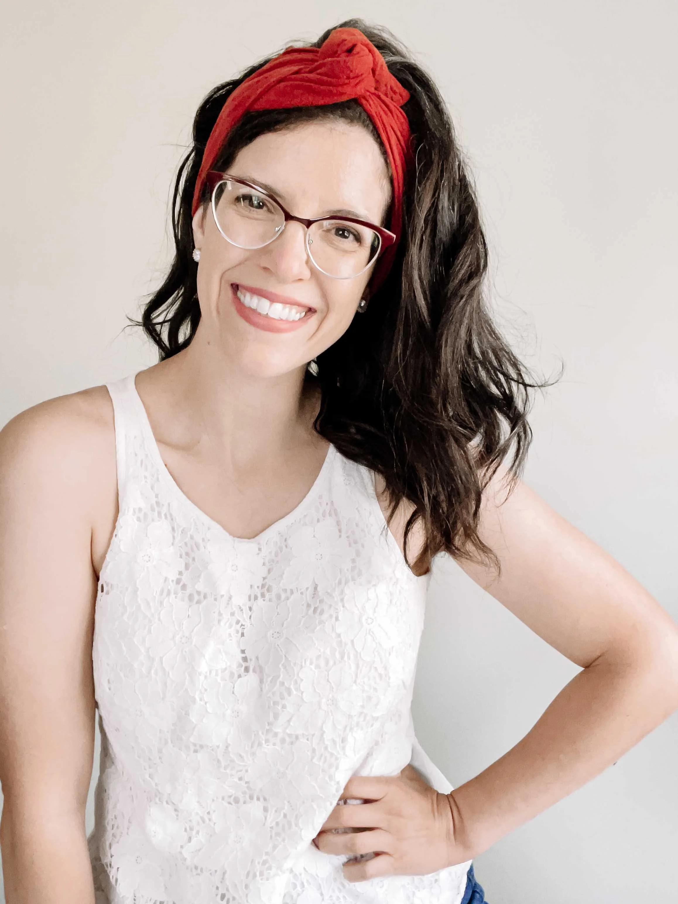 A woman with shoulder-length dark brown curly hair, wearing a red headband, glasses, a sleeveless white lace top, and earrings, smiling and standing with her left hand on her hip against a plain light-colored wall.