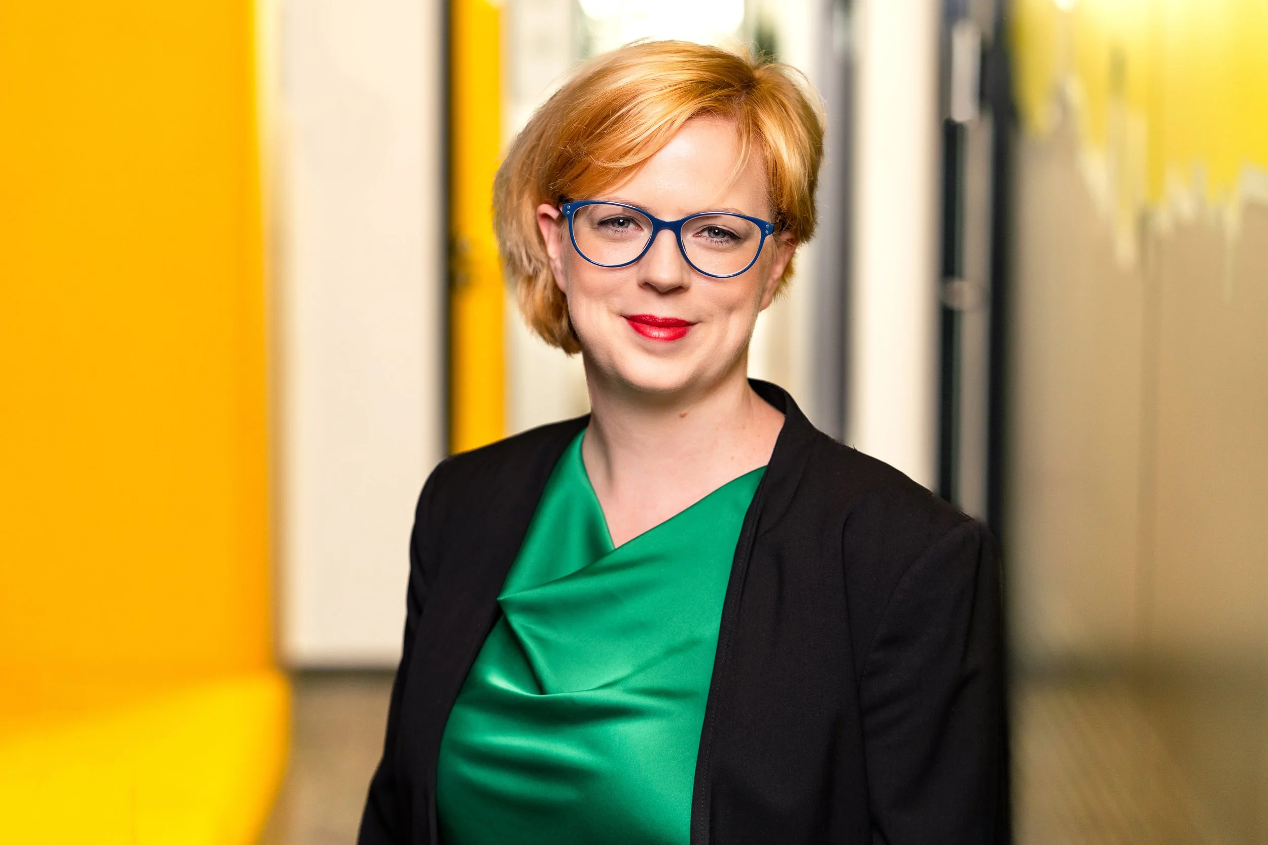 A woman with short blonde hair, wearing blue glasses, a green blouse, and a black blazer, standing in front of a yellow and white background.