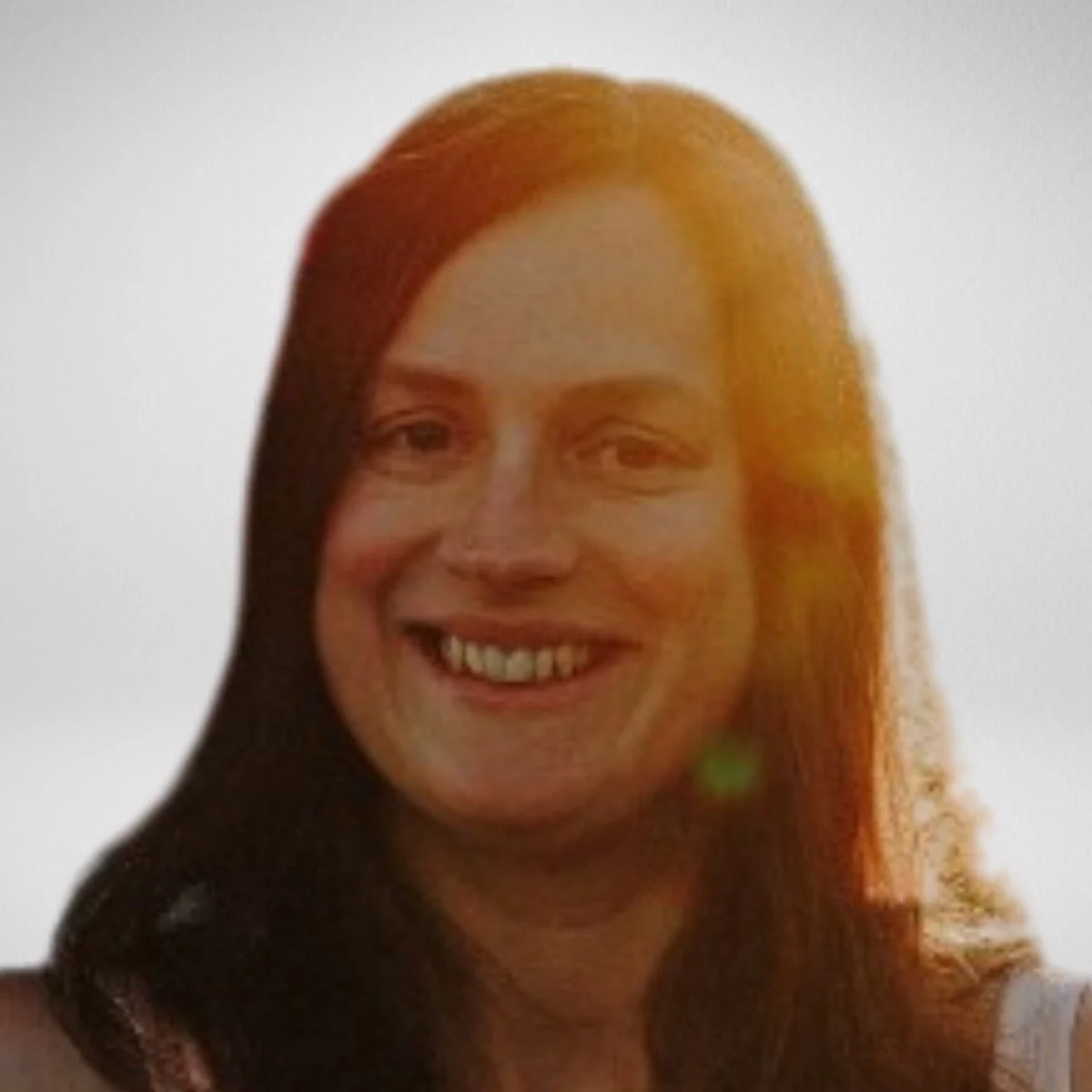 A woman with long reddish-brown hair smiling at the camera, with a bright background and sunlight flare.