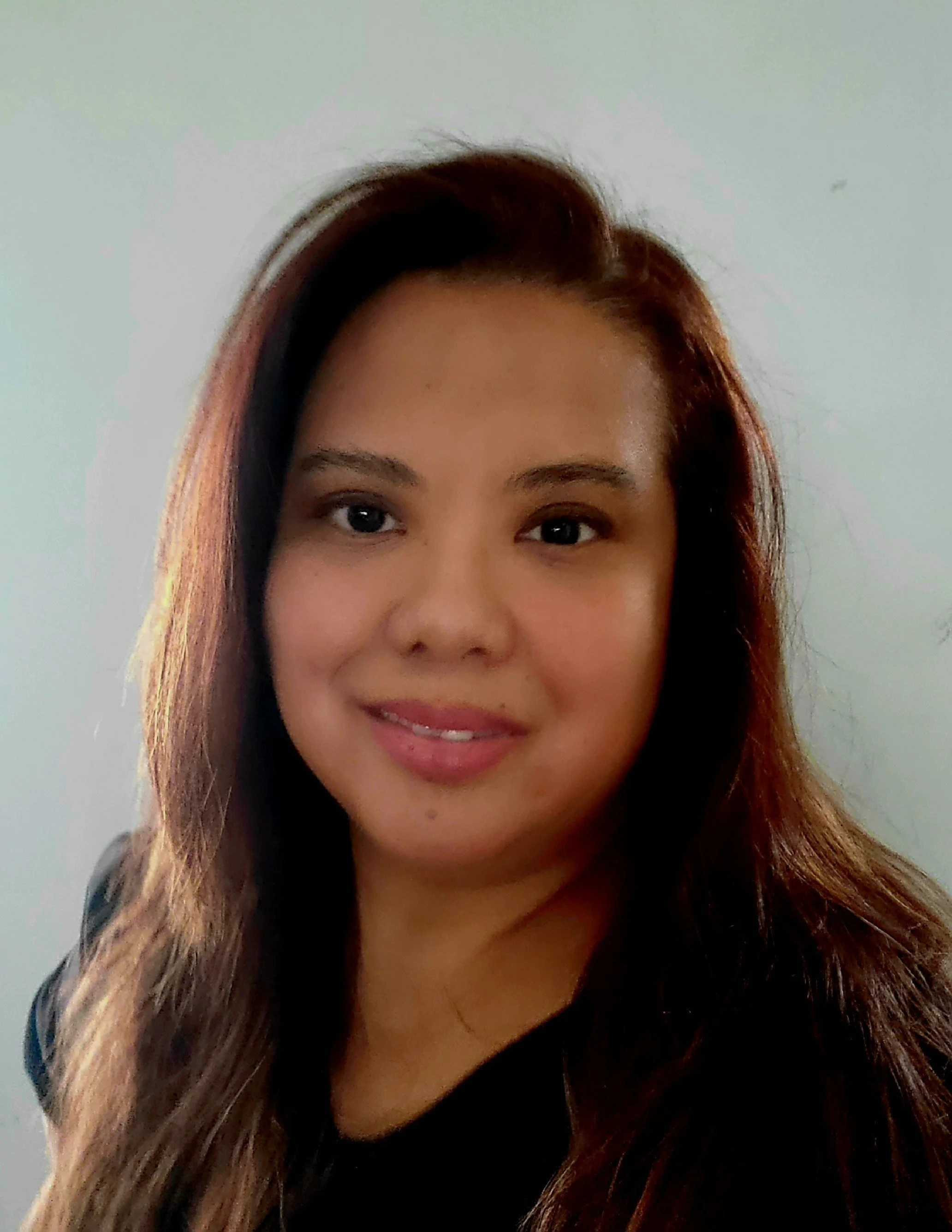 Portrait of a woman with long brown hair, smiling, against a plain light-colored background.
