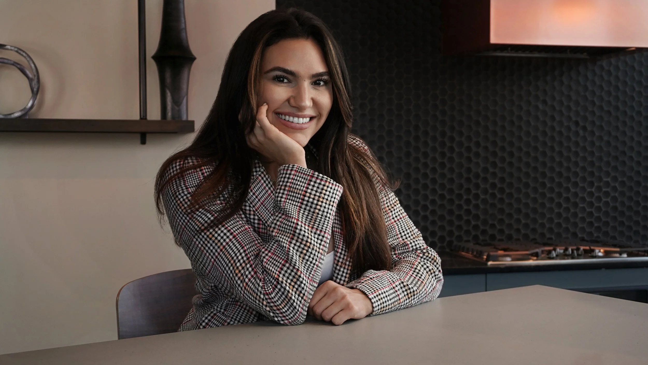 A woman with long brown hair smiling while sitting at a table in a modern kitchen.