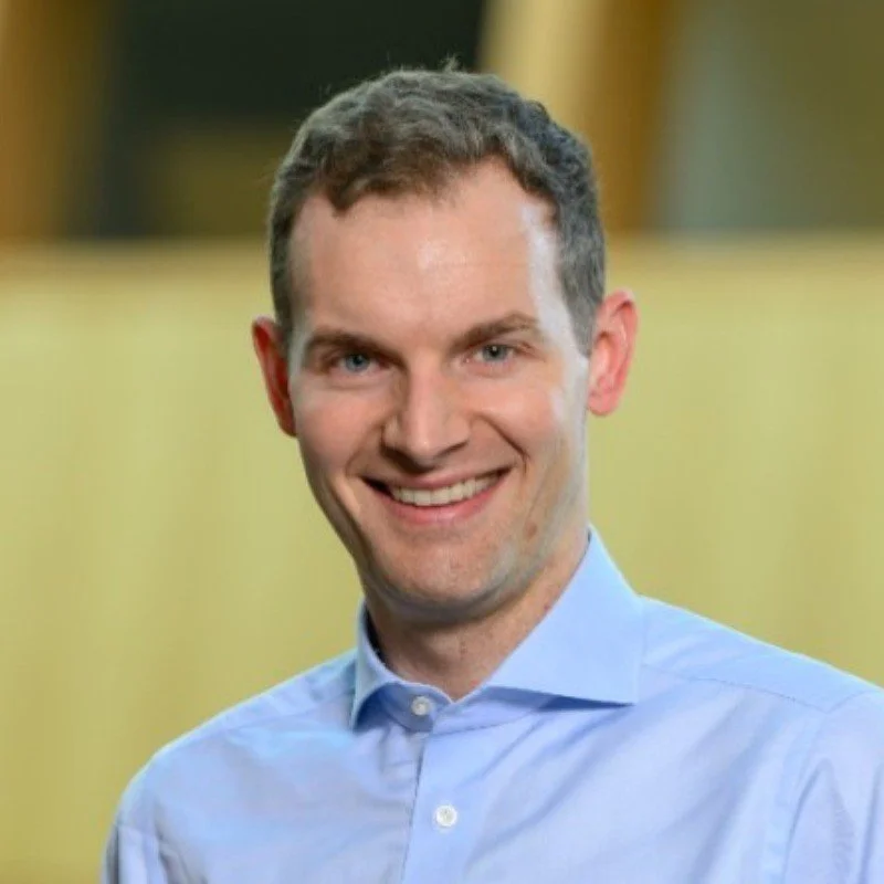 A young man with short brown hair wearing a light blue collared shirt, smiling at the camera.