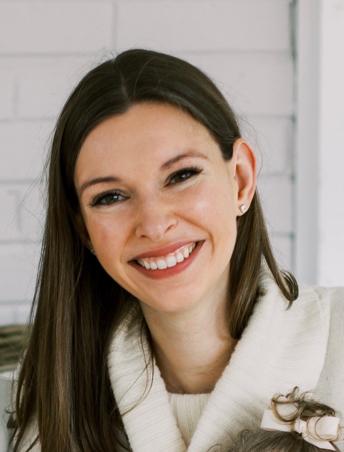 Close-up of a smiling woman with long brown hair, wearing a light-colored sweater, with a white brick wall in the background.