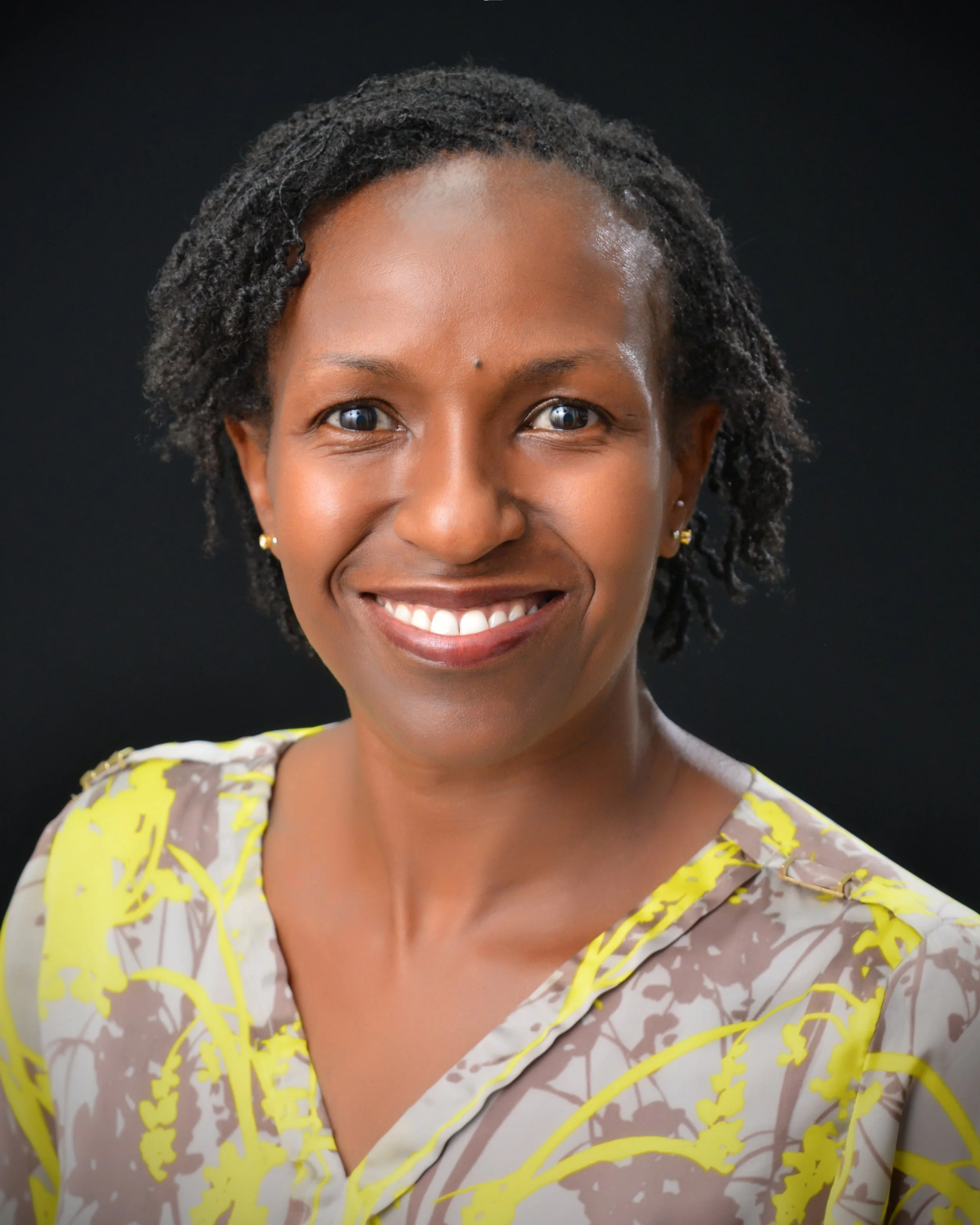 A smiling african american woman with short curly hair wearing a yellow and gray patterned blouse, against a black background.