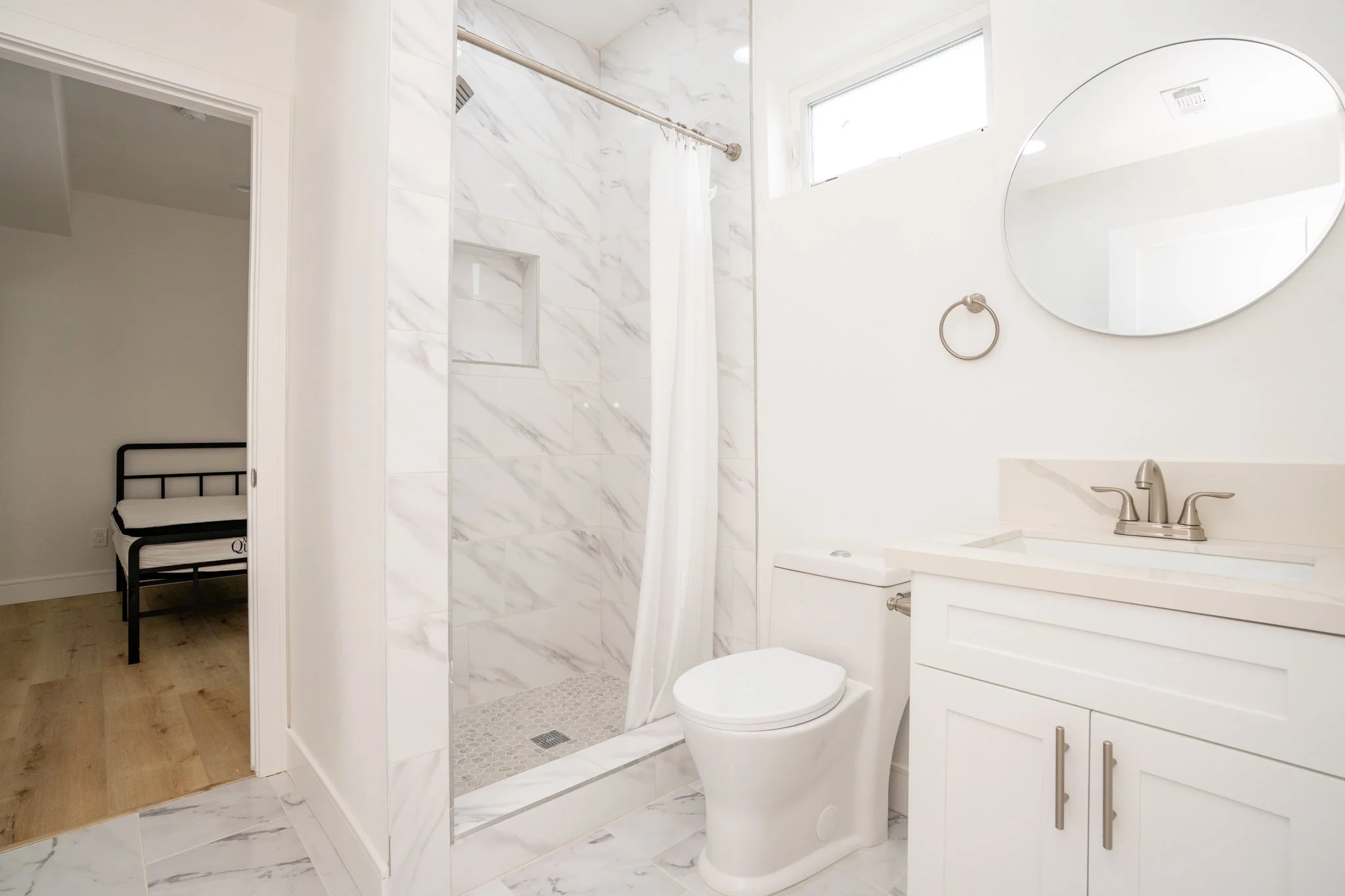 Modern bathroom with white marble tiles, a shower with curtain, toilet, white vanity with sink, and round mirror. Visible glimpse of an adjoining room with a bed frame.