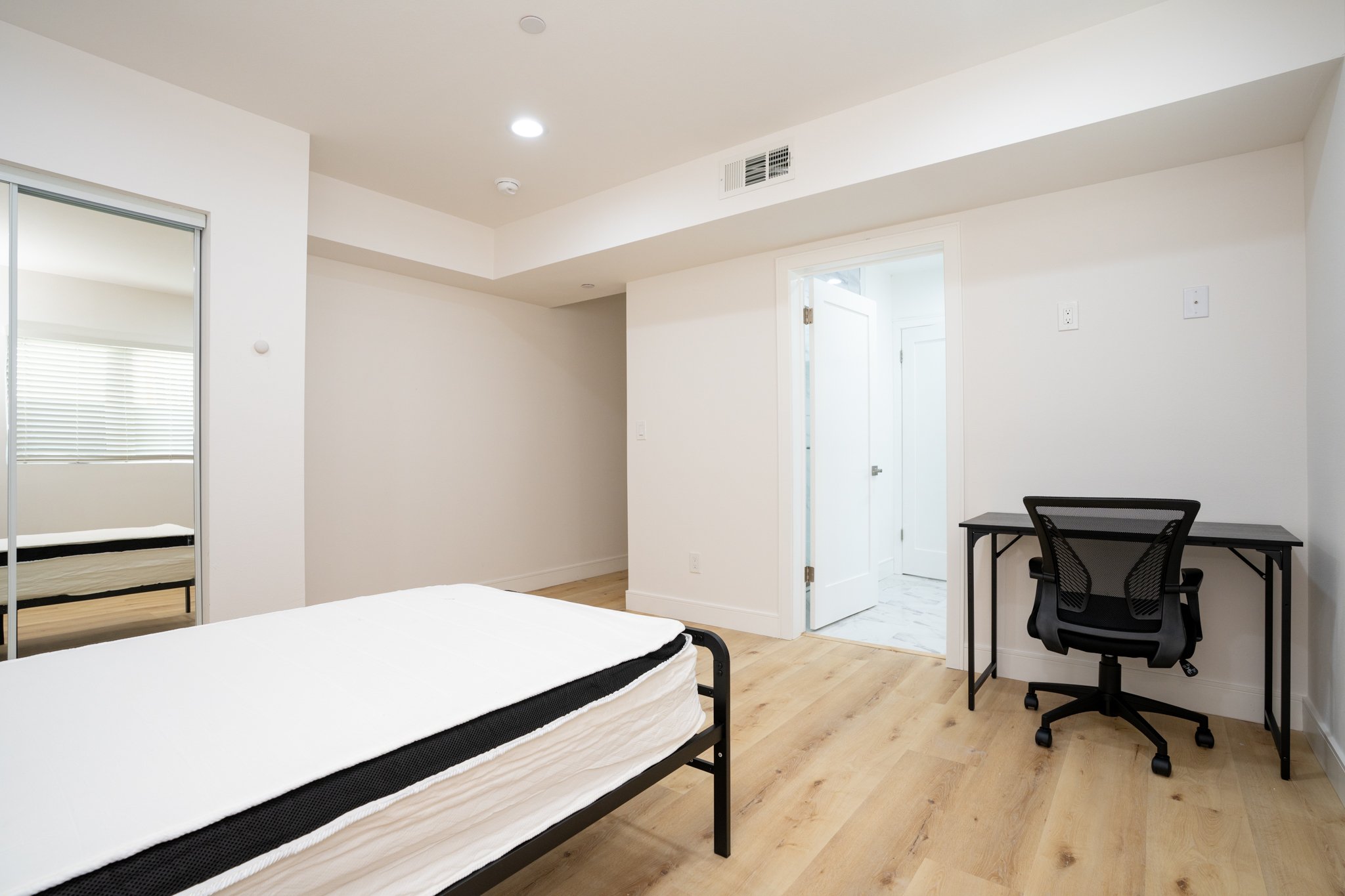 Minimalist bedroom with wooden floor, single bed with white bedding, desk with black chair, mirrored closet, and door leading to bathroom.