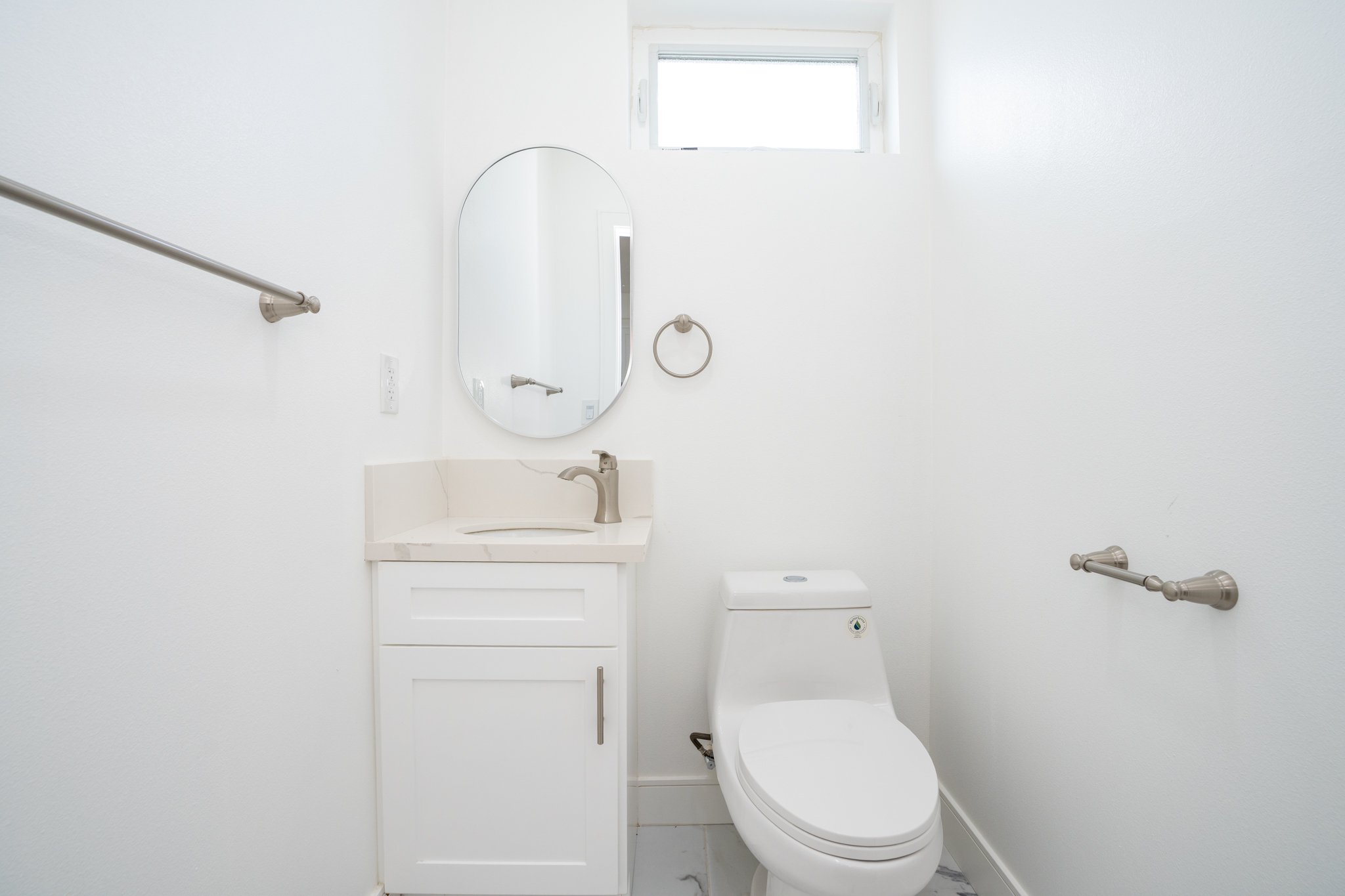 Minimalist bathroom with a white toilet, vanity with a sink, oval mirror, and stainless steel fixtures.