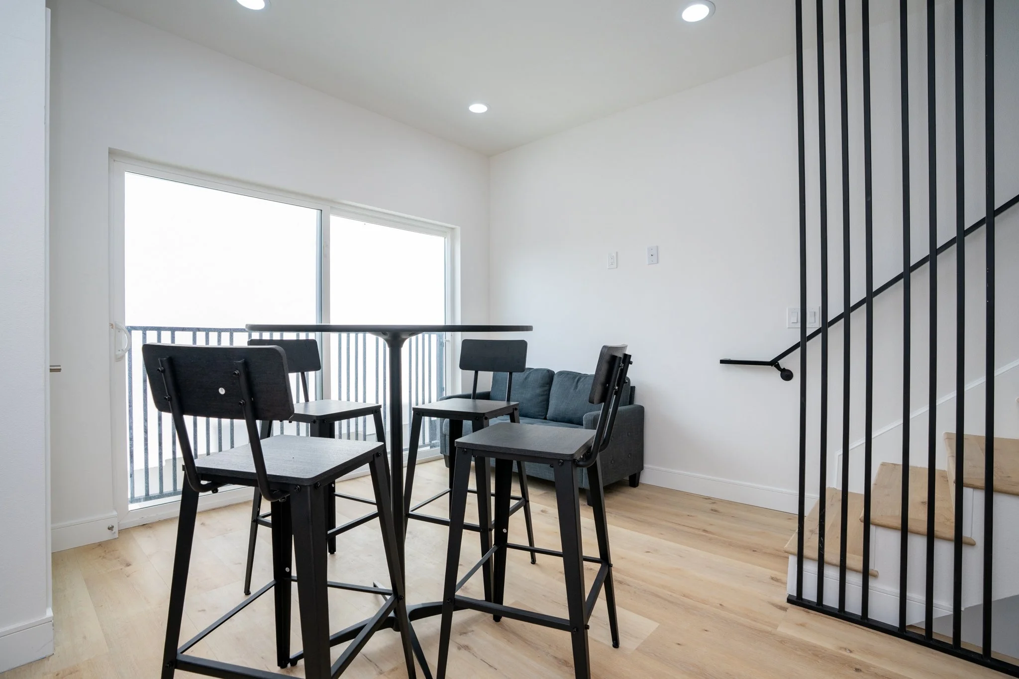 Modern dining area with high chairs, round table, and gray sofa near stairs with metal railings, large window.
