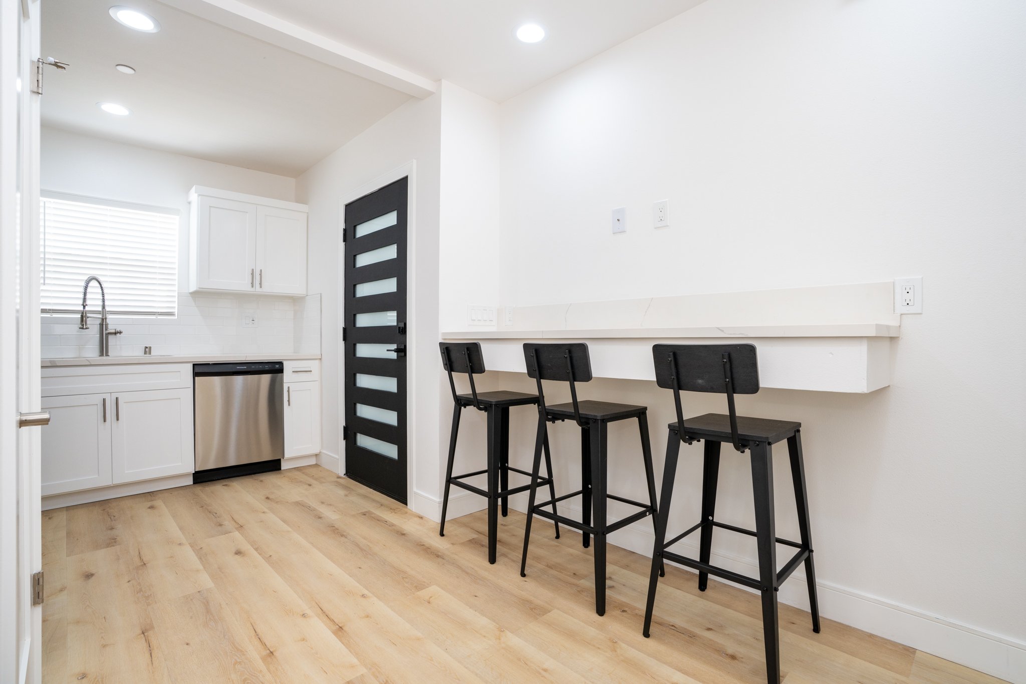 Minimalistic kitchen with white cabinets, stainless steel appliances, bar counter with three black stools, and light wood flooring.