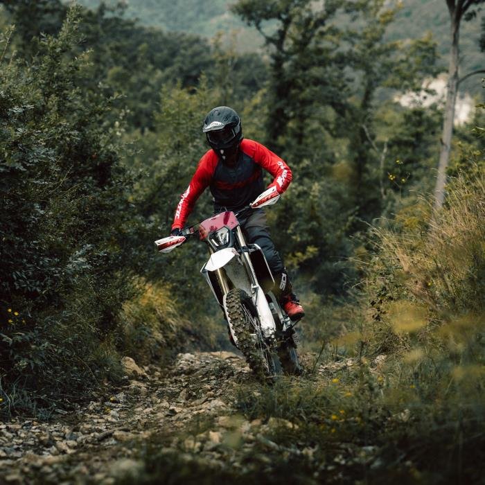 A motorcyclist wearing a helmet and red protective gear riding a dirt bike on a rocky forest trail.
