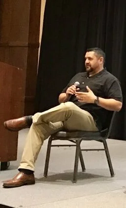 Man sitting on a chair holding a microphone, wearing a black shirt and beige pants.