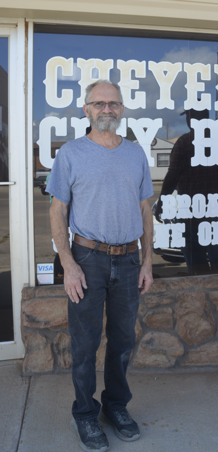 An elderly man with glasses and a beard standing outside a store with large lettering on the window, wearing a blue t-shirt, dark jeans, and sneakers.