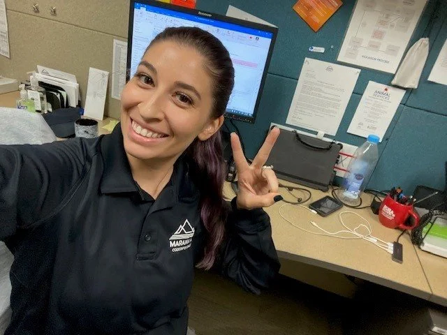 A woman smiling and making a peace sign in an office cubicle with a computer and various office supplies.