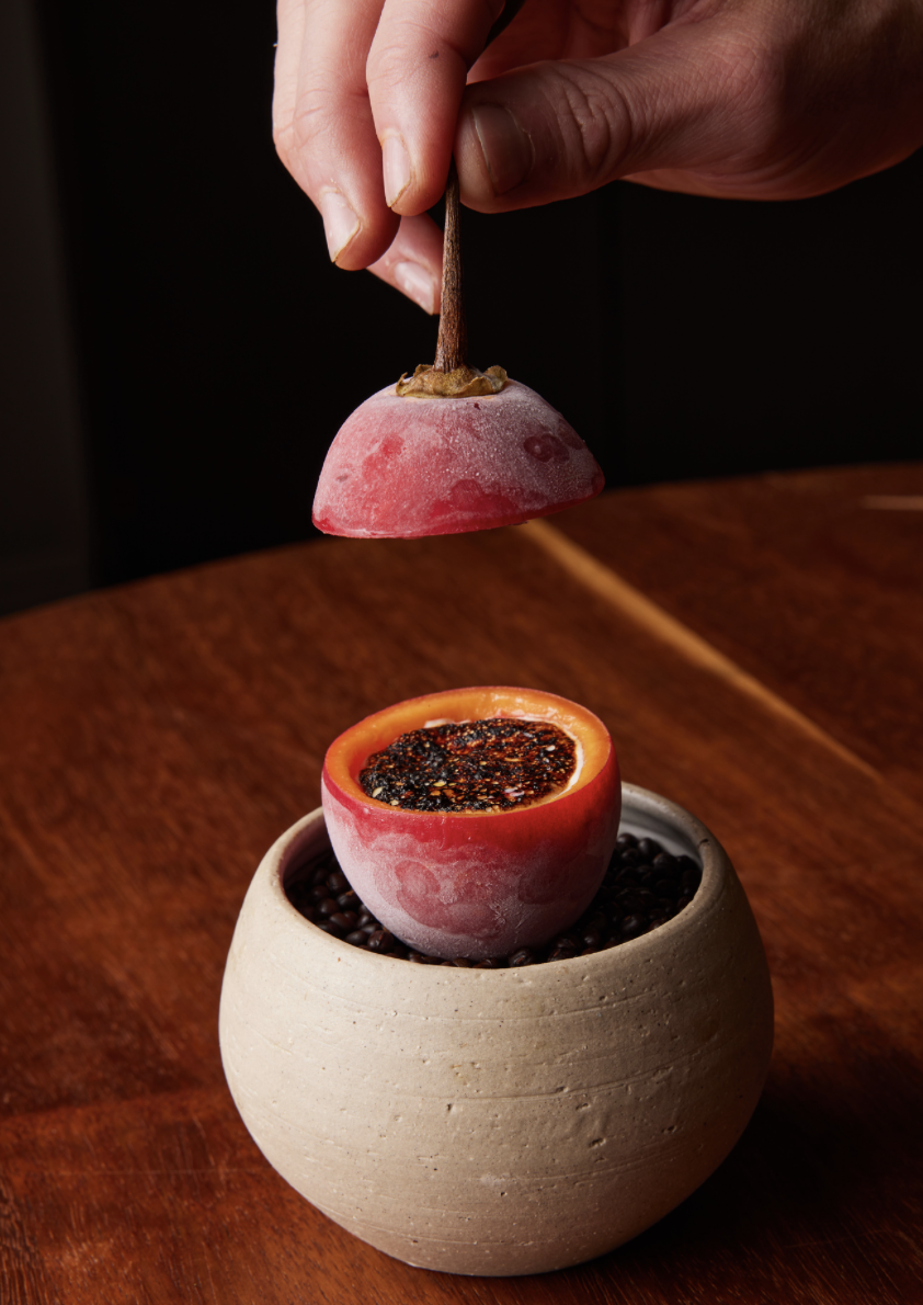 Chef Sid Sahrawat's Frozen Tamarillo with seeds, held by a hand, placed in a bowl with black beans on a wooden table.