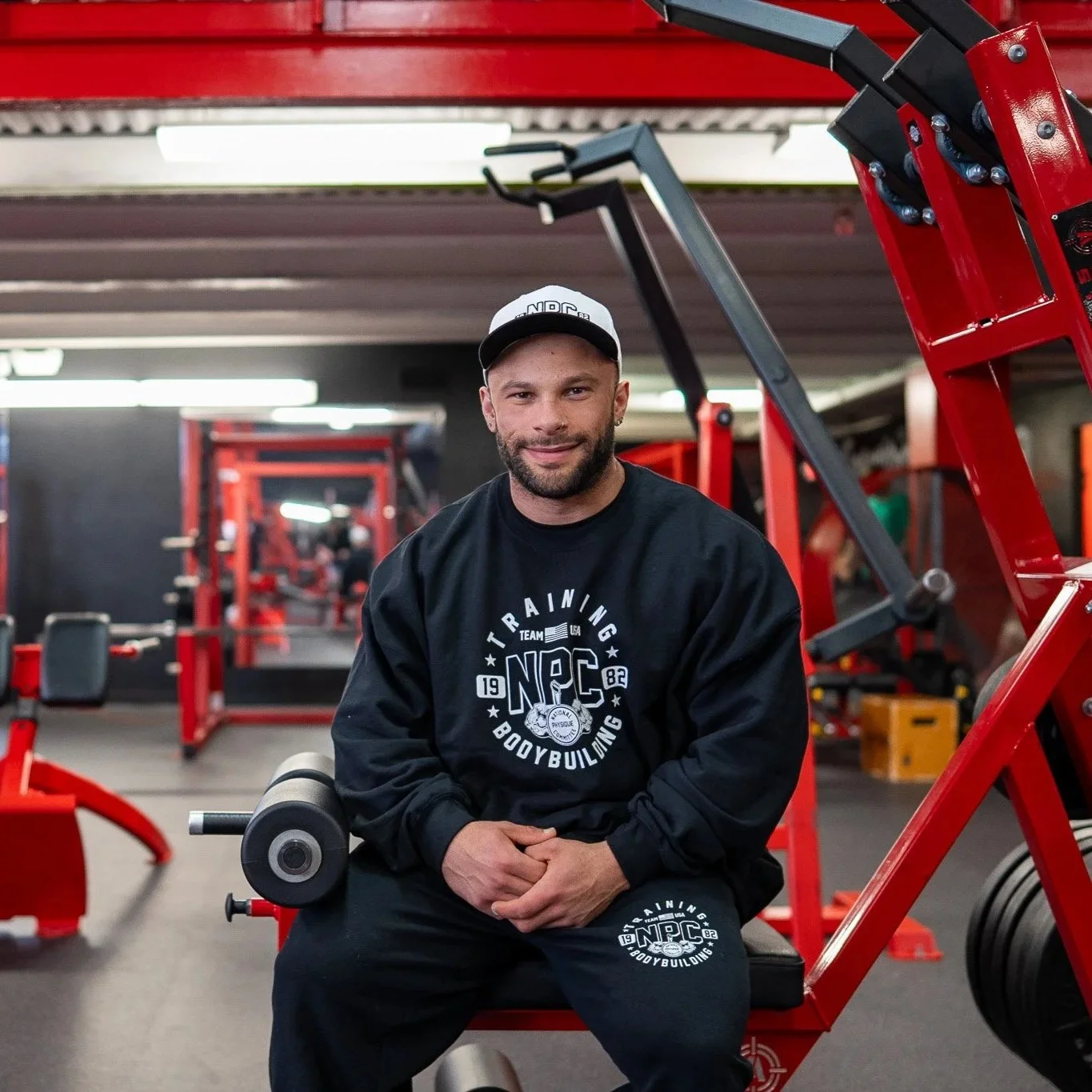 A man sitting on gym equipment in a fitness center, wearing a black sweatshirt and sweatpants with 'NPC Training Bodybuilding' logo, and a white cap.