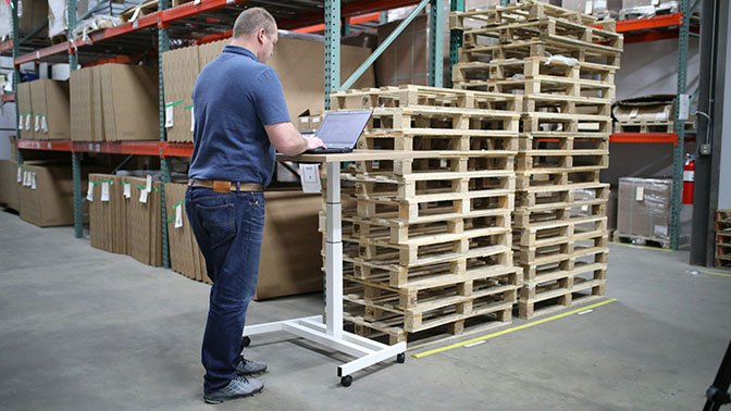 A man in a warehouse using a laptop on a standing desk next to stacked wooden pallets.