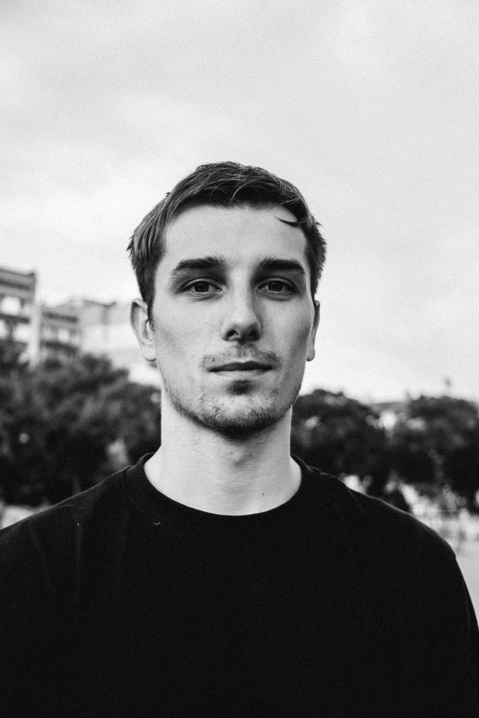 Black and white portrait of a young man with short hair, wearing a dark shirt, standing outdoors with blurred trees and buildings in the background.