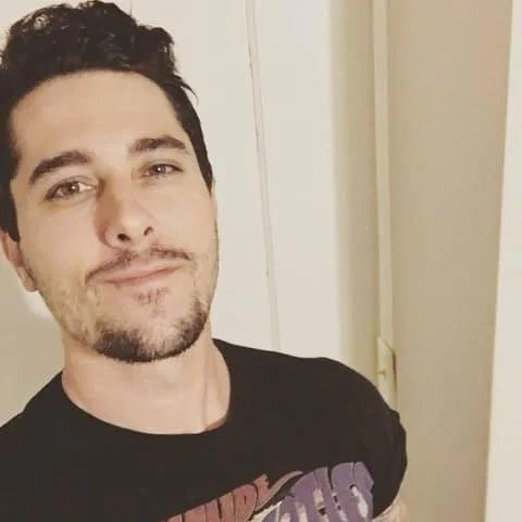 A young man with dark hair, beard, and mustache taking a selfie indoors, wearing a black T-shirt with partially visible text.